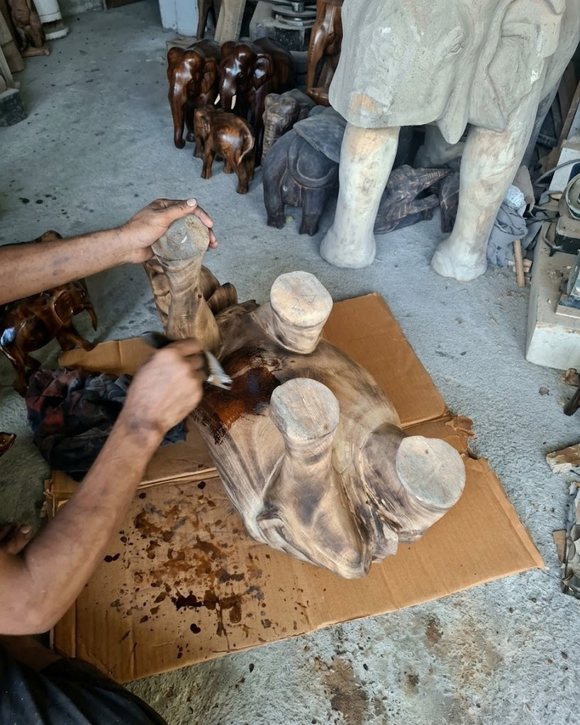 A hand brushing a dark finish onto a large carved elephant laid on cardboard, finished carvings standing on the shelves behind.