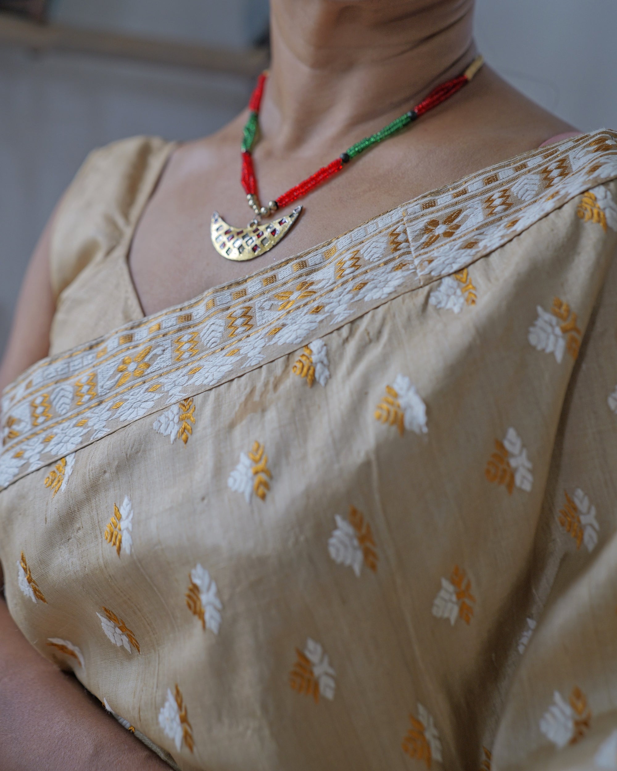 At the neckline, the ivory and golden-orange border crossing the collarbone, small two-tone motifs below it, worn with a red and green bead necklace and a crescent pendant.