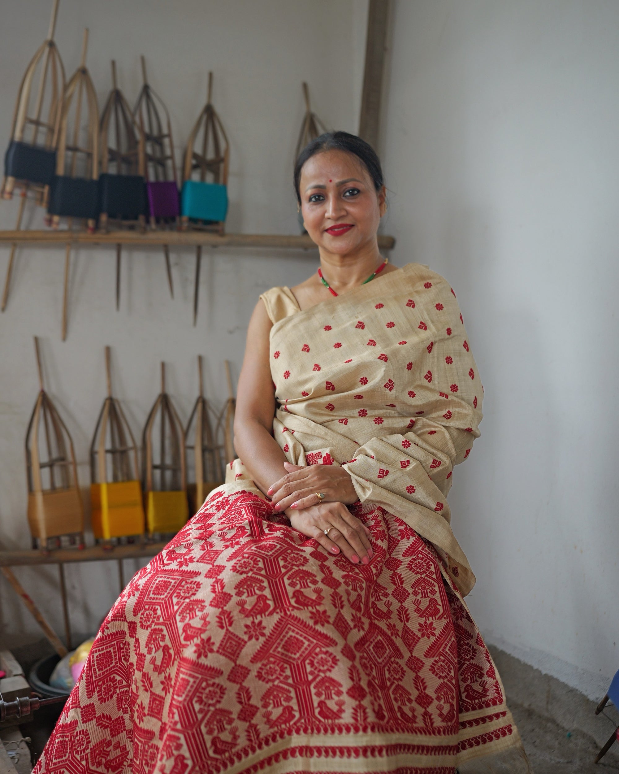 Seated beside a wall of bamboo reels wound with coloured yarn, smiling. Small red motifs scatter the pale gold drape over the shoulder, and the dense red field of the pallu spreads across the lap to the floor.