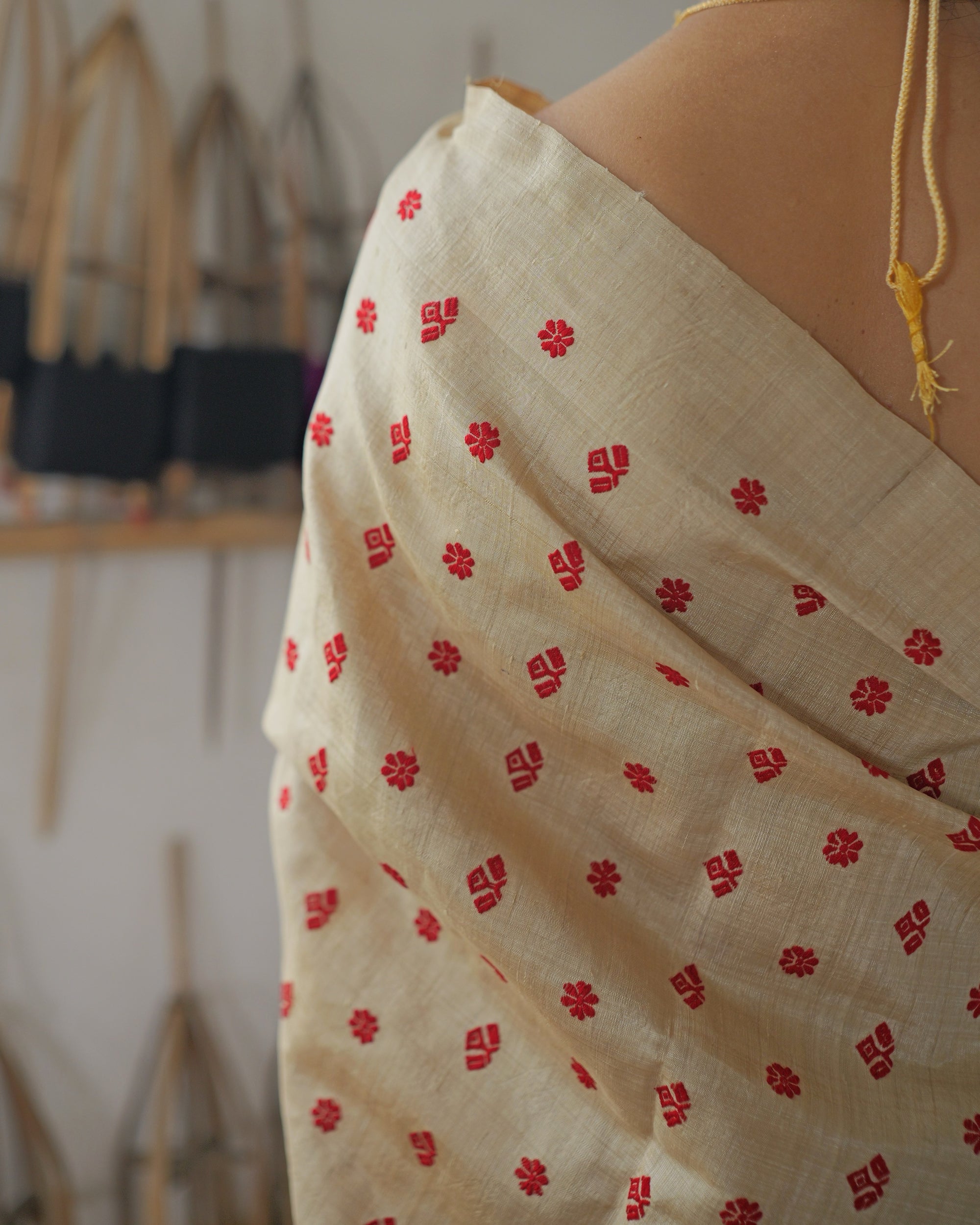 Over the shoulder from behind, a gold cord hanging at the back of the blouse, small red motifs running down the gold drape, bamboo reels soft behind.