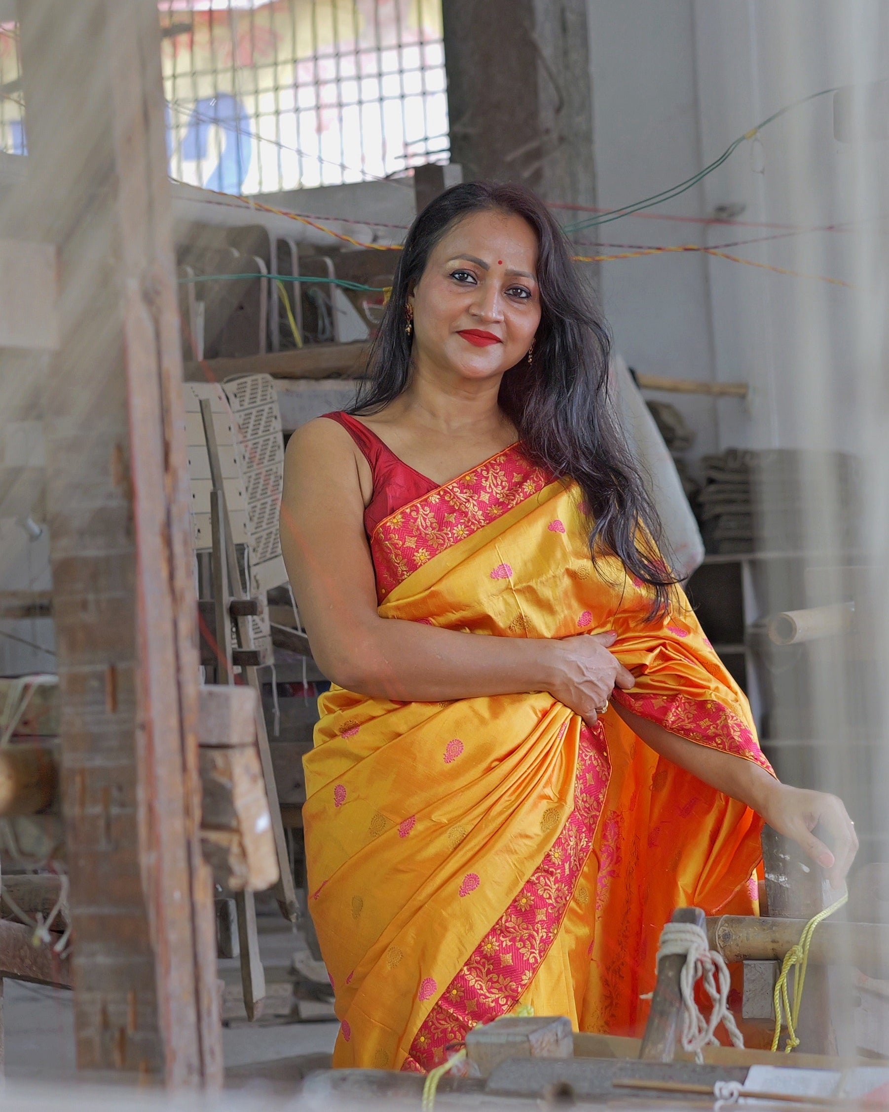 A loom shed seen through the timber frame of a loom, a marigold and pink saree lit by the window beyond it.