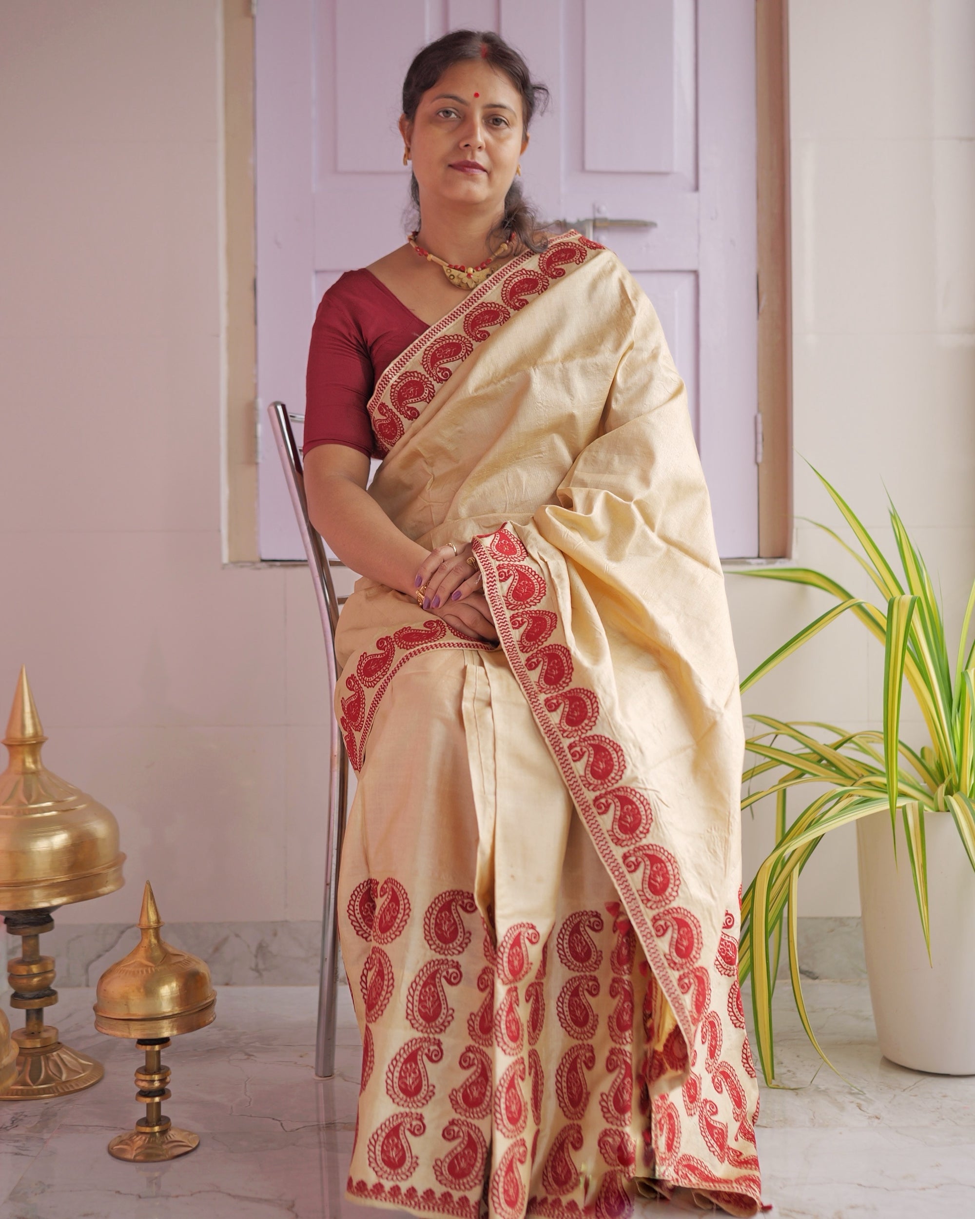 Seated on a chair against a panelled door, the whole set visible. A plain golden-cream body with a border and hem of large red paisleys, worn with a maroon blouse.