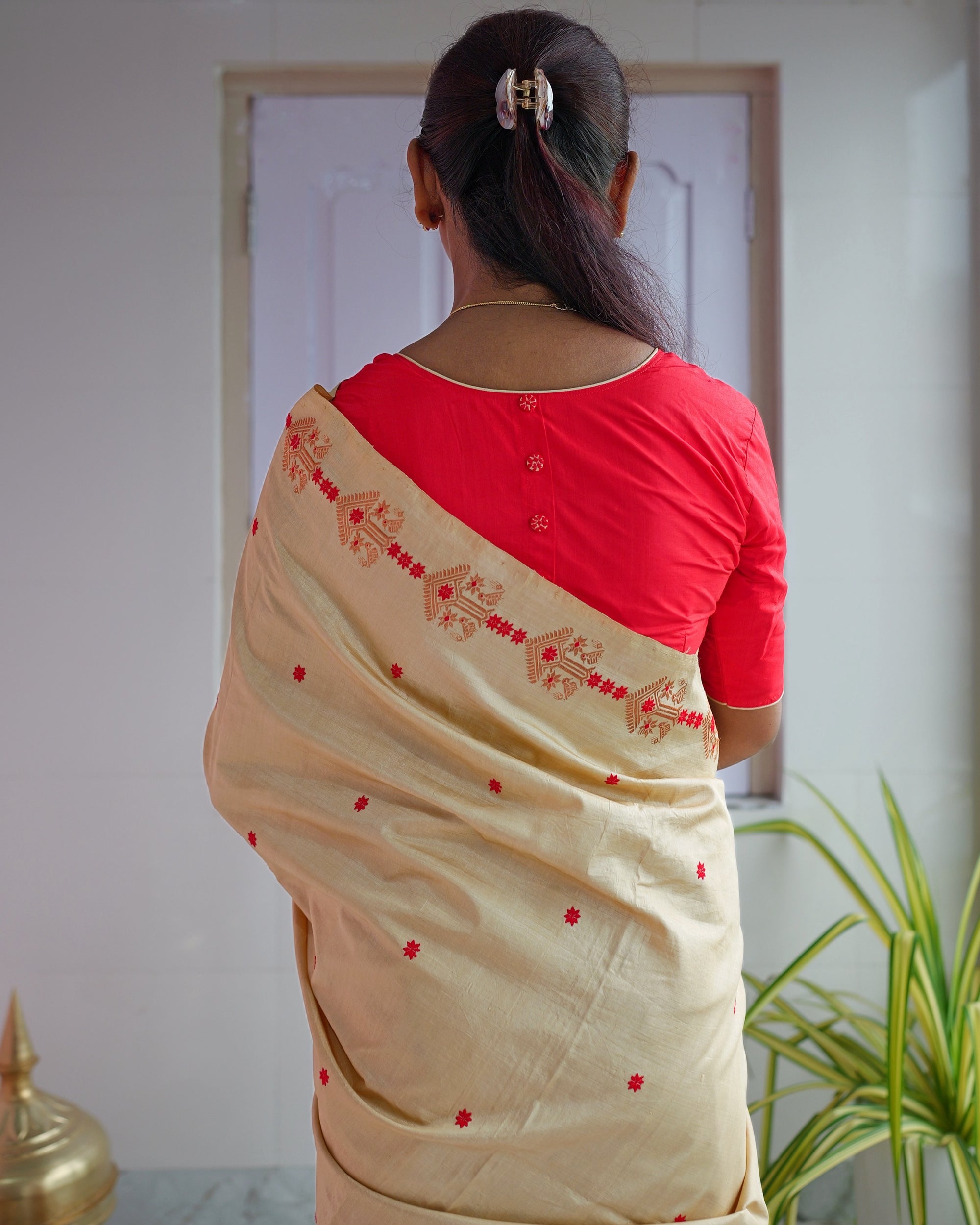 Seen from behind in a red blouse, hair held up in a clip. The drape falls over the shoulder and down the back, a line of copper-peach motifs along its edge and small red florets across it.