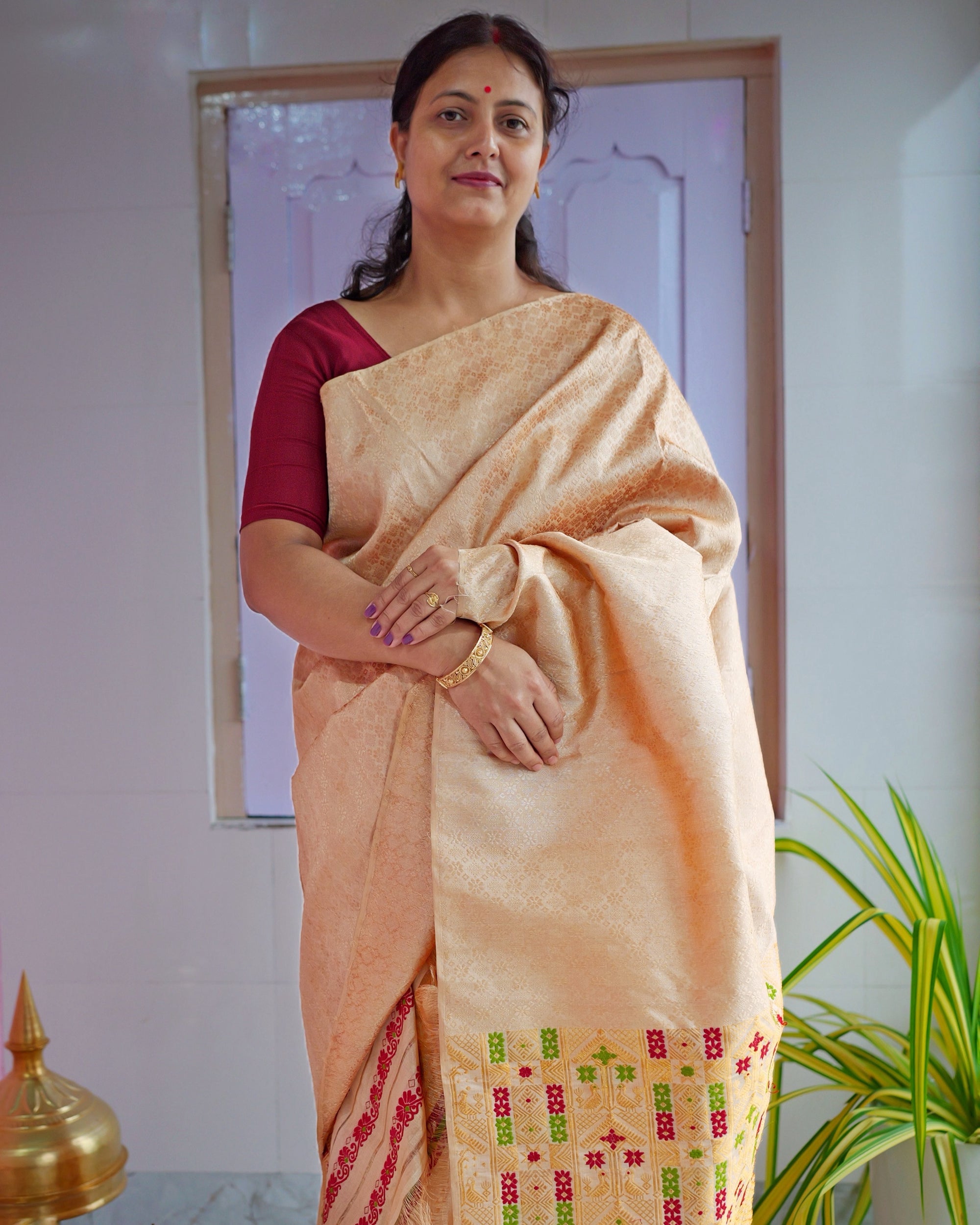 Standing against a lilac panelled door in a maroon blouse, hands folded at the waist, framed from head to knee. A golden-peach drape with a faint tonal pattern throughout, its end worked in red and green at the lower right.