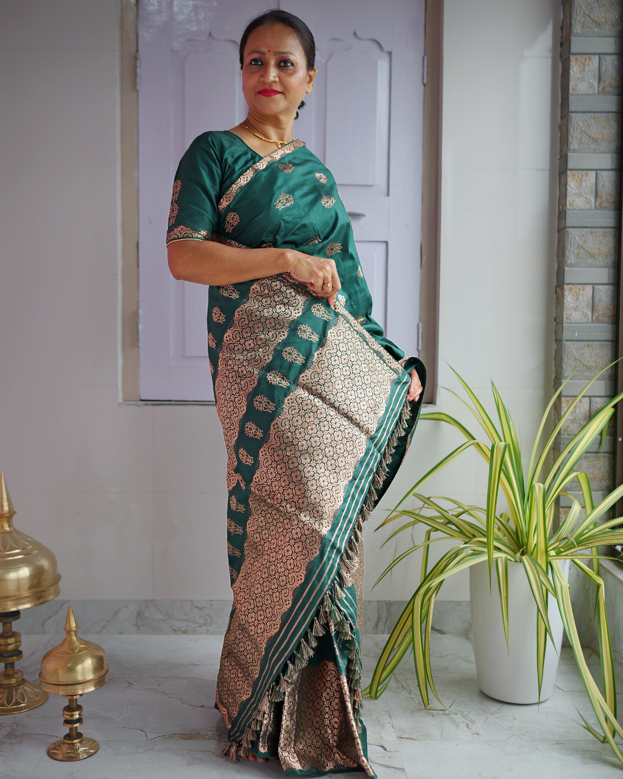 Standing against a panelled door, the whole set visible. A deep emerald body scattered with rose-gold buti, over a hem panel of dense zari lattice.