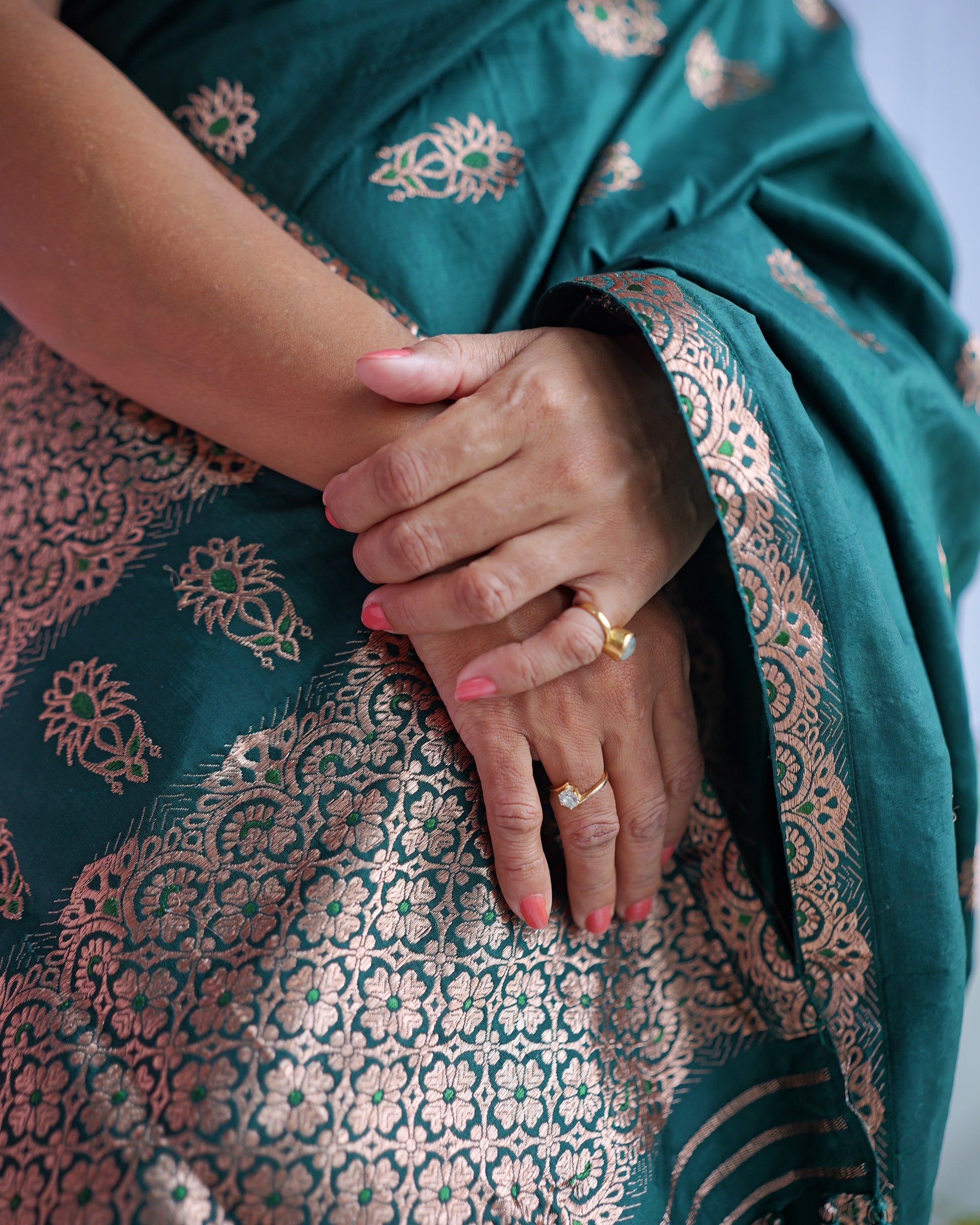 Hands resting over the drape, rings on the fingers. Rose-gold buti on emerald, above the dense zari lattice of the hem.