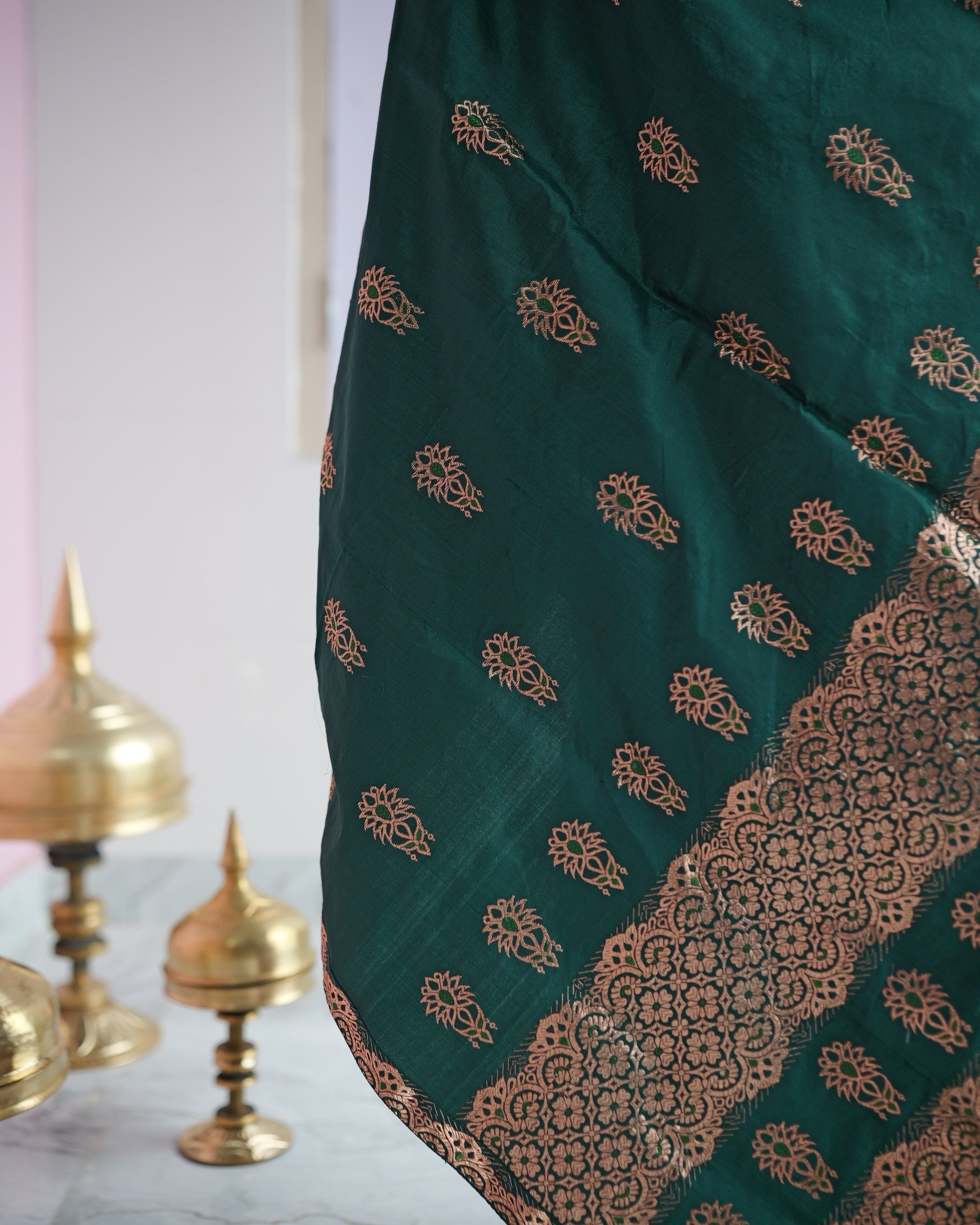 The emerald drape hanging in front of brass xorai on the floor, rows of rose-gold buti above a band of rosette lattice with a scalloped edge.