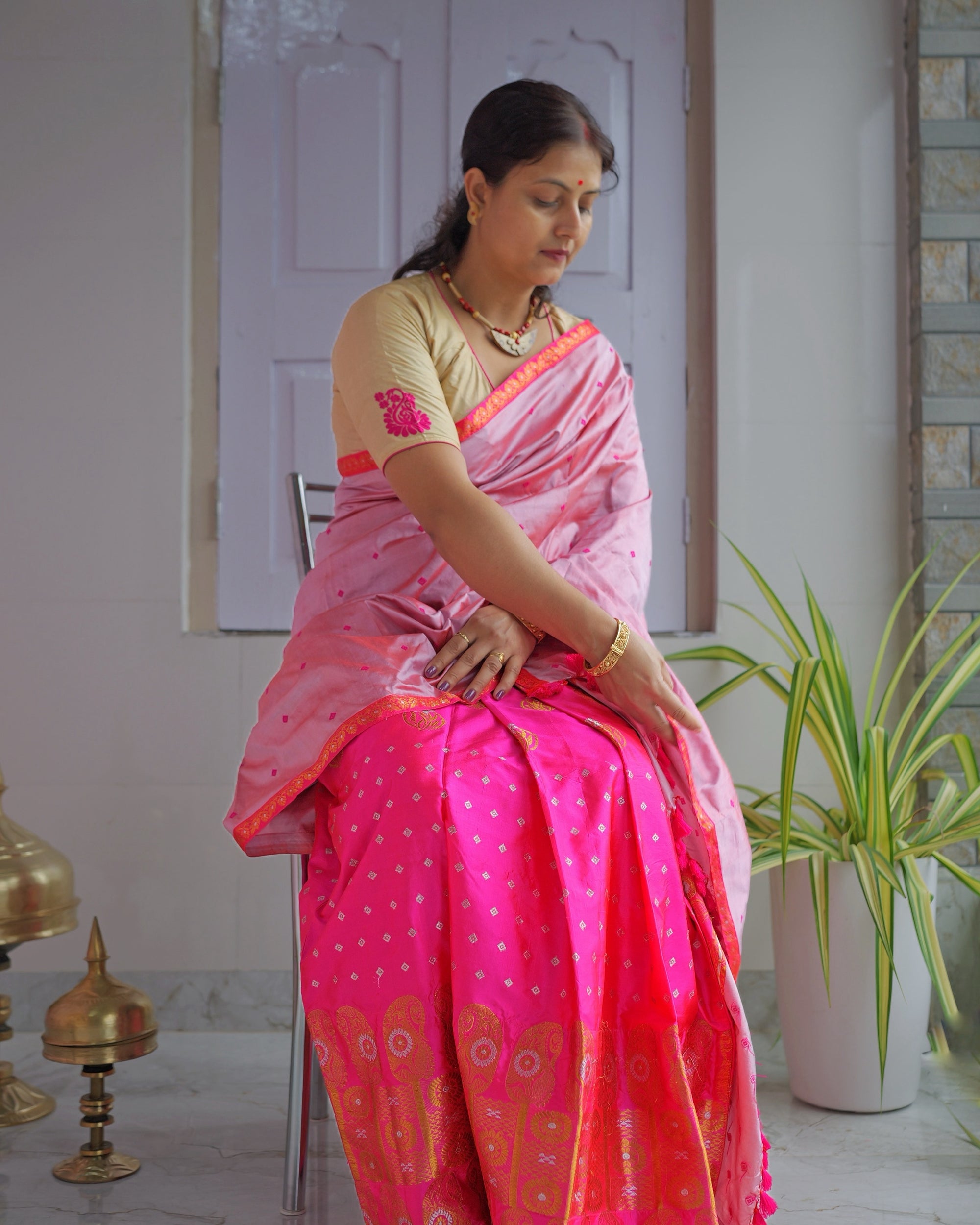 Seated on a chair against a panelled door, the whole set visible. A rose-pink drape over a vivid magenta mekhela worked with golden-orange zari at the hem.