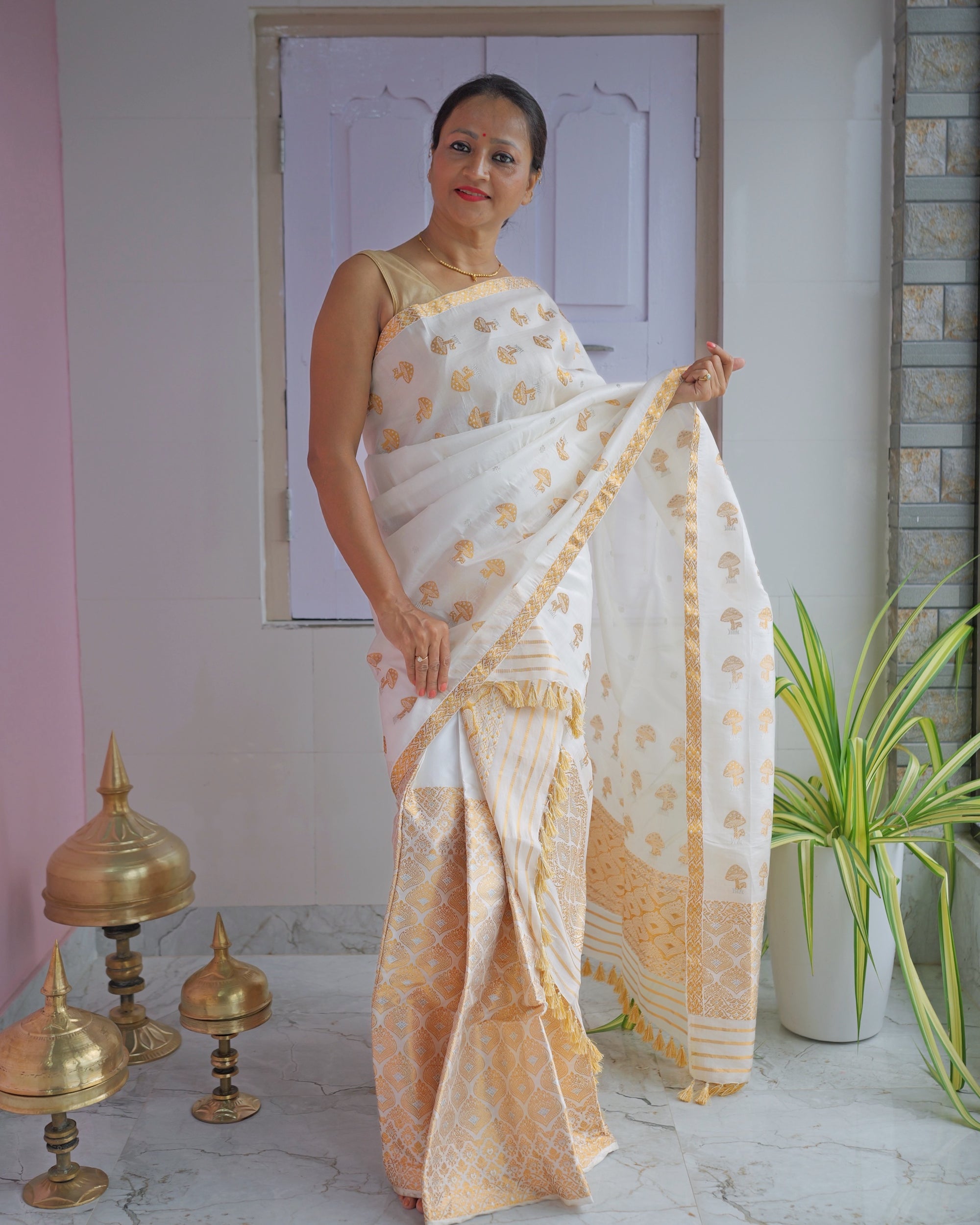 Standing against a panelled door, the whole set visible. An ivory body scattered with small gold mushroom motifs, over a mekhela hem worked in dense gold zari.
