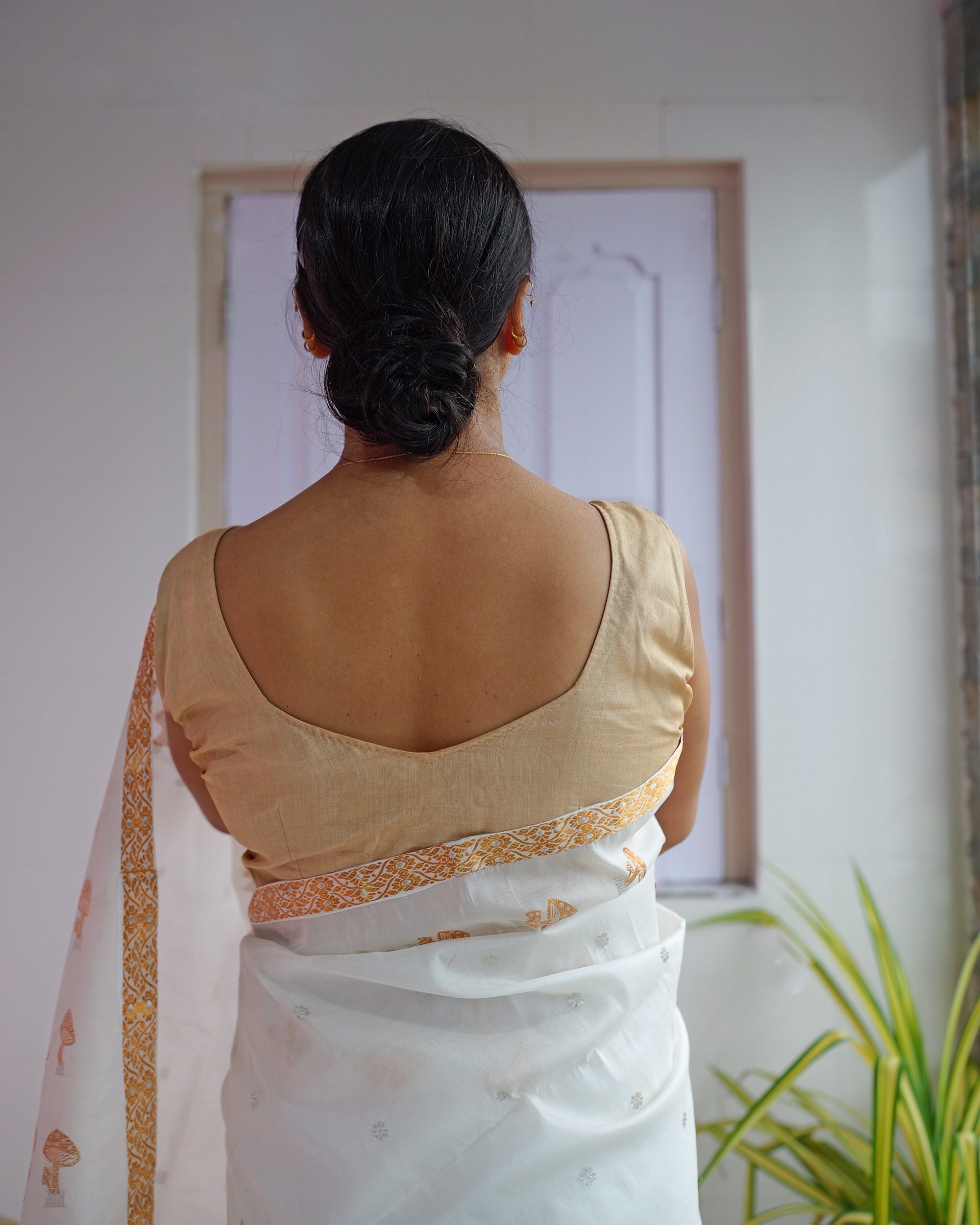 Seen from behind, a sleeveless gold-beige blouse above the ivory drape: a zari border wraps the waist, small silver-toned florets dot the cloth below it, and the loose end of the sador hangs at one side.