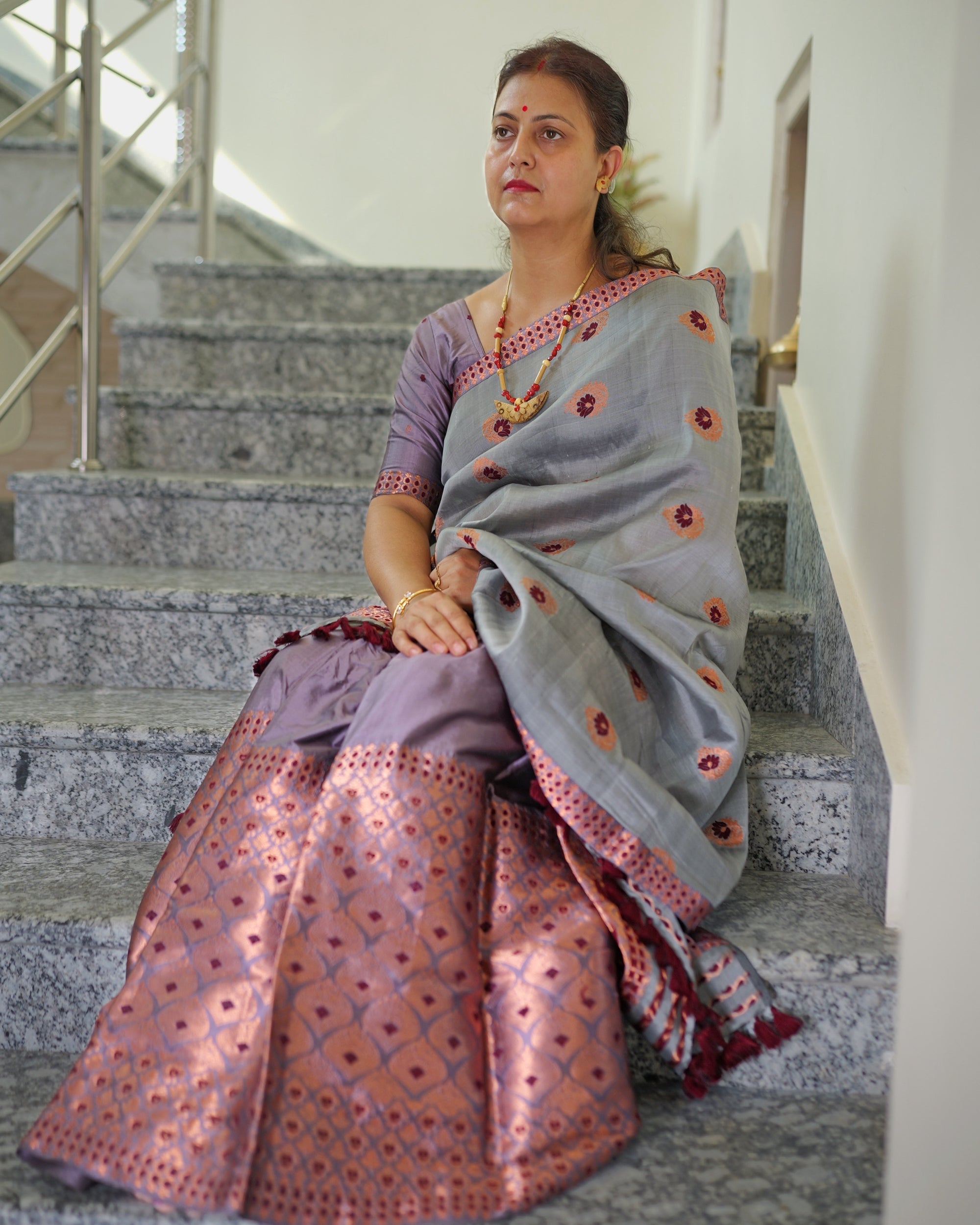 Seated on a granite staircase, looking off to one side, the whole set visible. A grey drape scattered with round maroon and copper flowers, over a lilac mekhela whose lower half is copper lattice, maroon tassels at the sador end.
