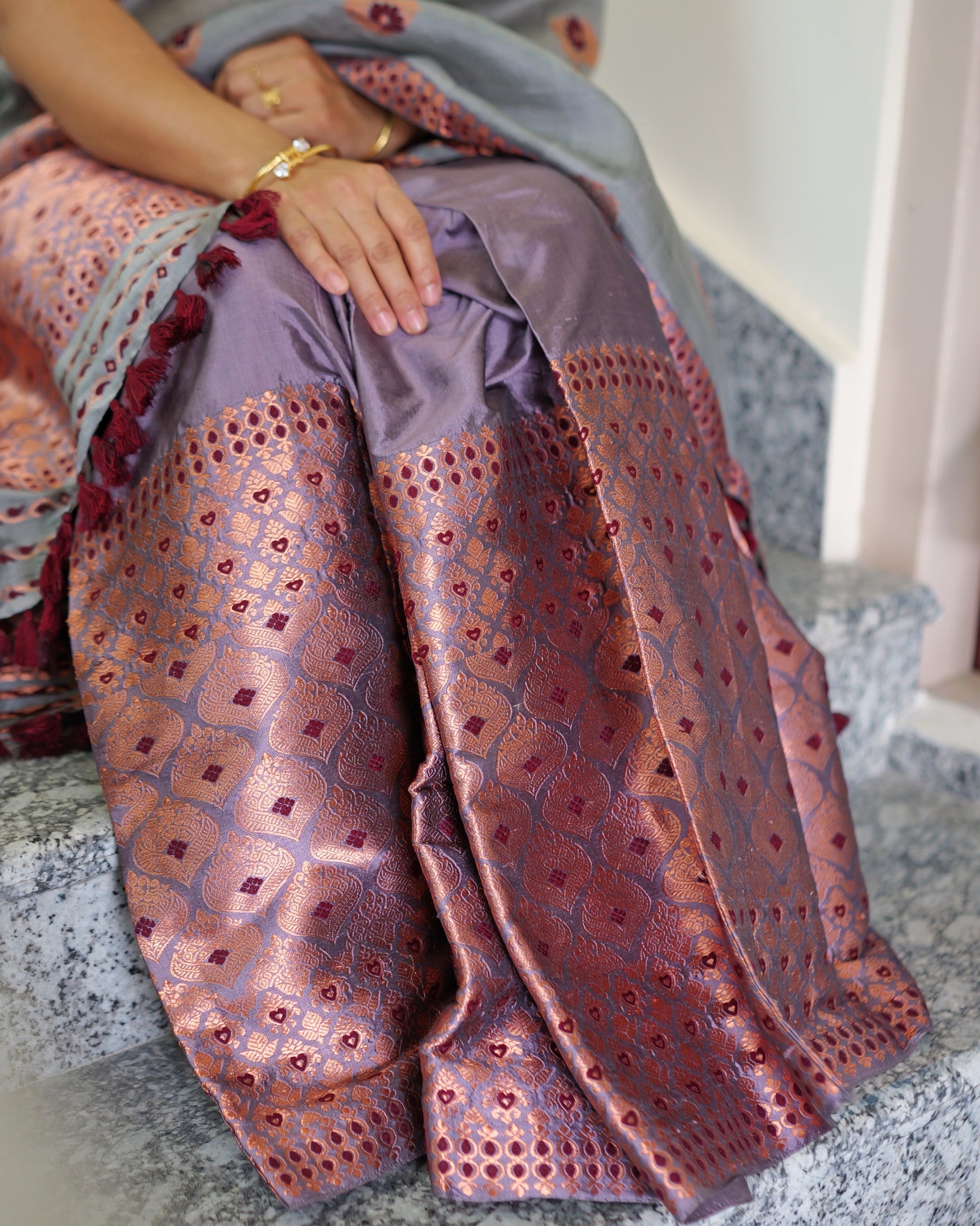 Hands with rings and a gold bangle resting on the lilac mekhela, its copper lattice falling over the stair, the grey sador and its maroon tassels at one side.