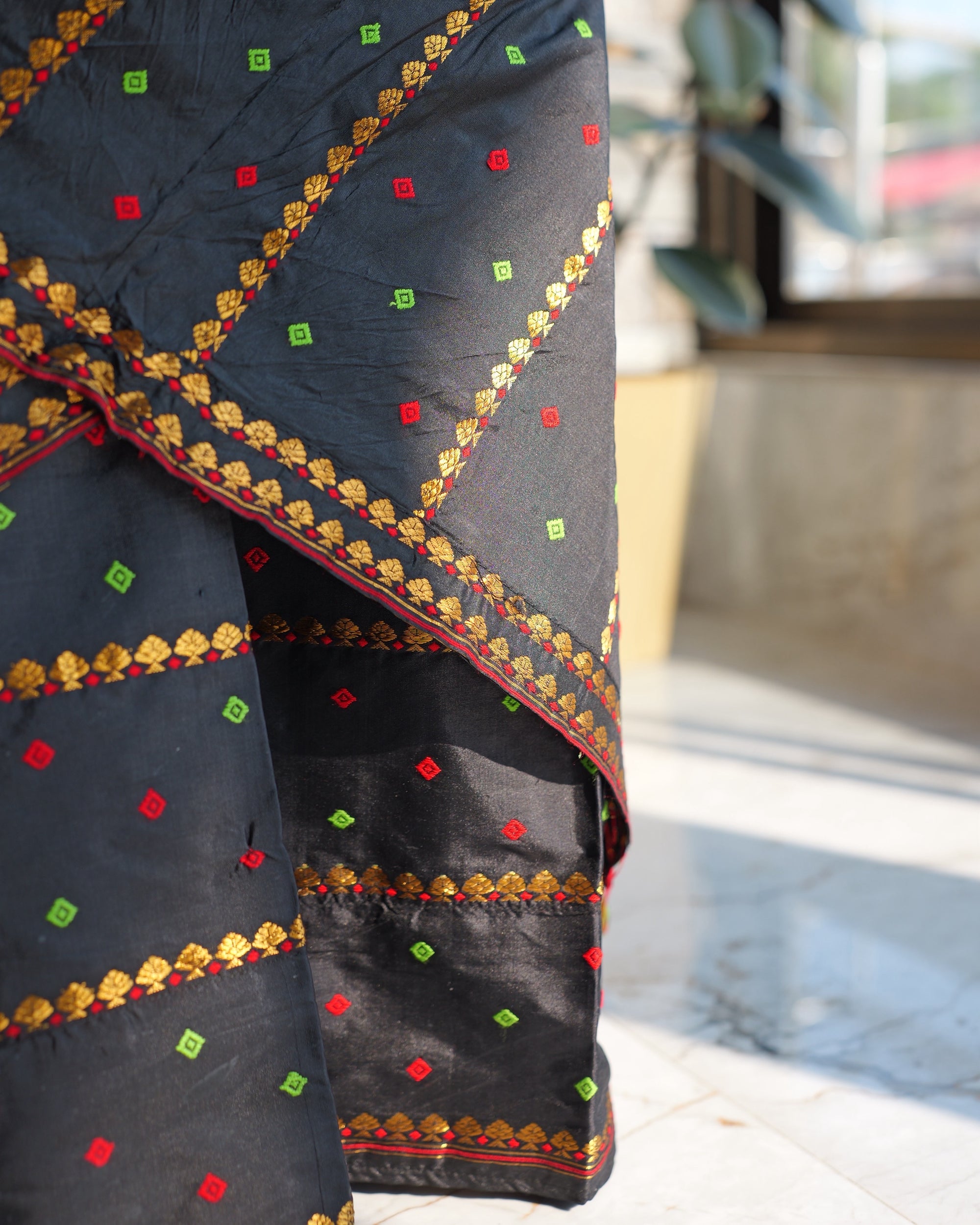 The sador's border close up where it crosses the mekhela: a row of small gold fan shapes tipped with red and traced by a fine red line, red and green squares on the black.