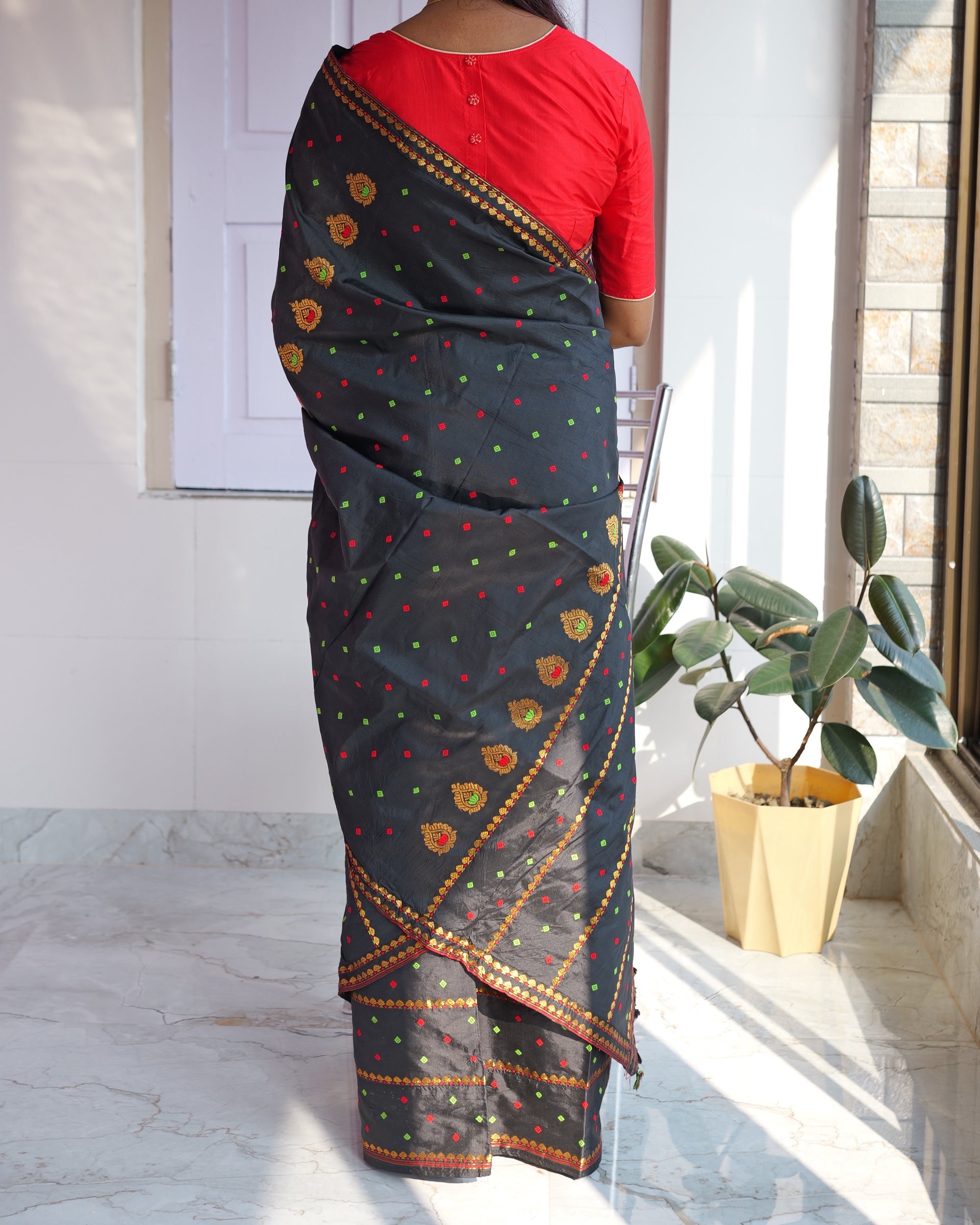 Seen from behind, standing, from the shoulders down: the black sador falling down the back over a red blouse, round gold flowers along it and a gold border running diagonally to the hem.