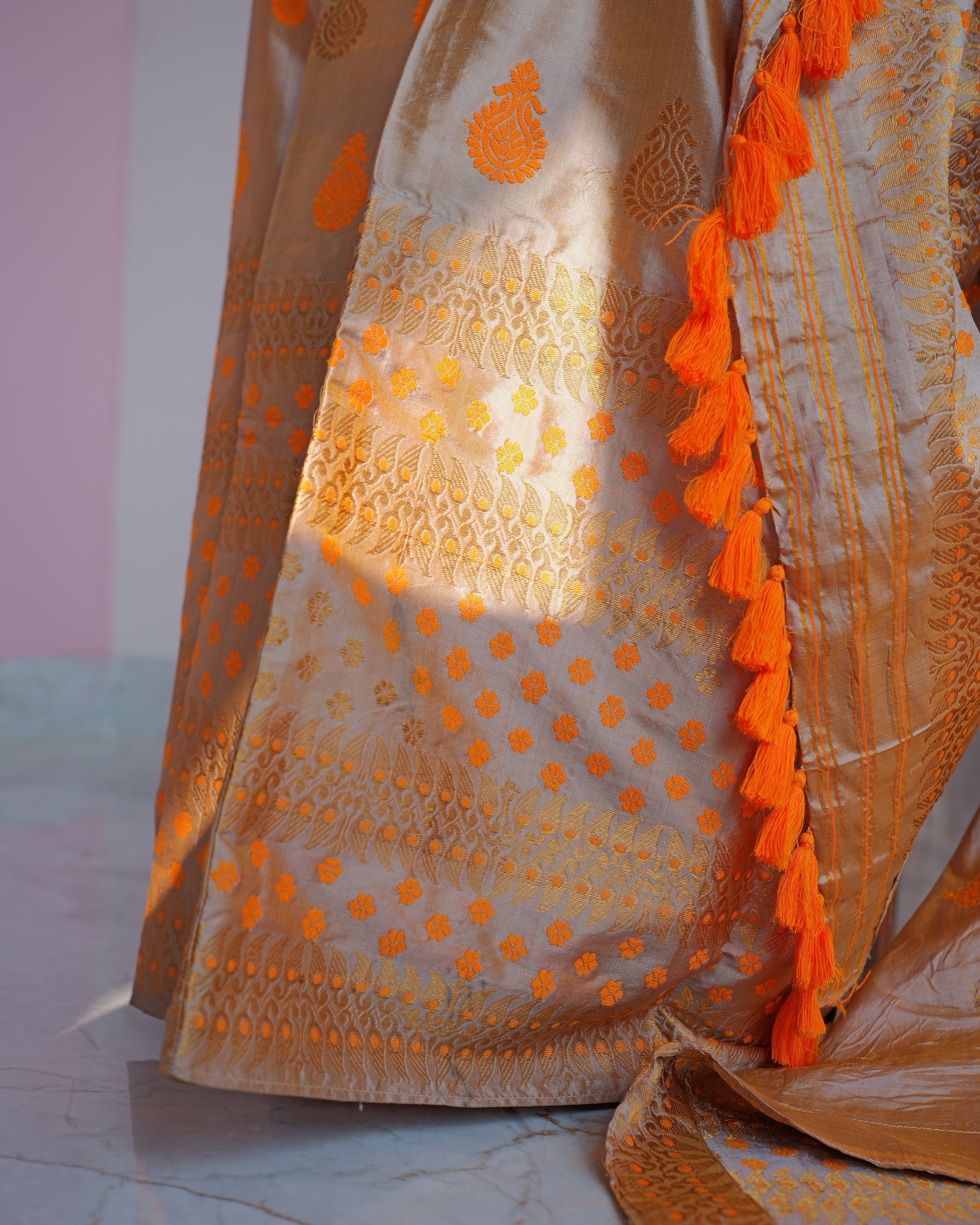 The sador end over the mekhela in a patch of sun: bands of gold-toned zari dotted with saffron florets, and a line of saffron tassels down the edge.
