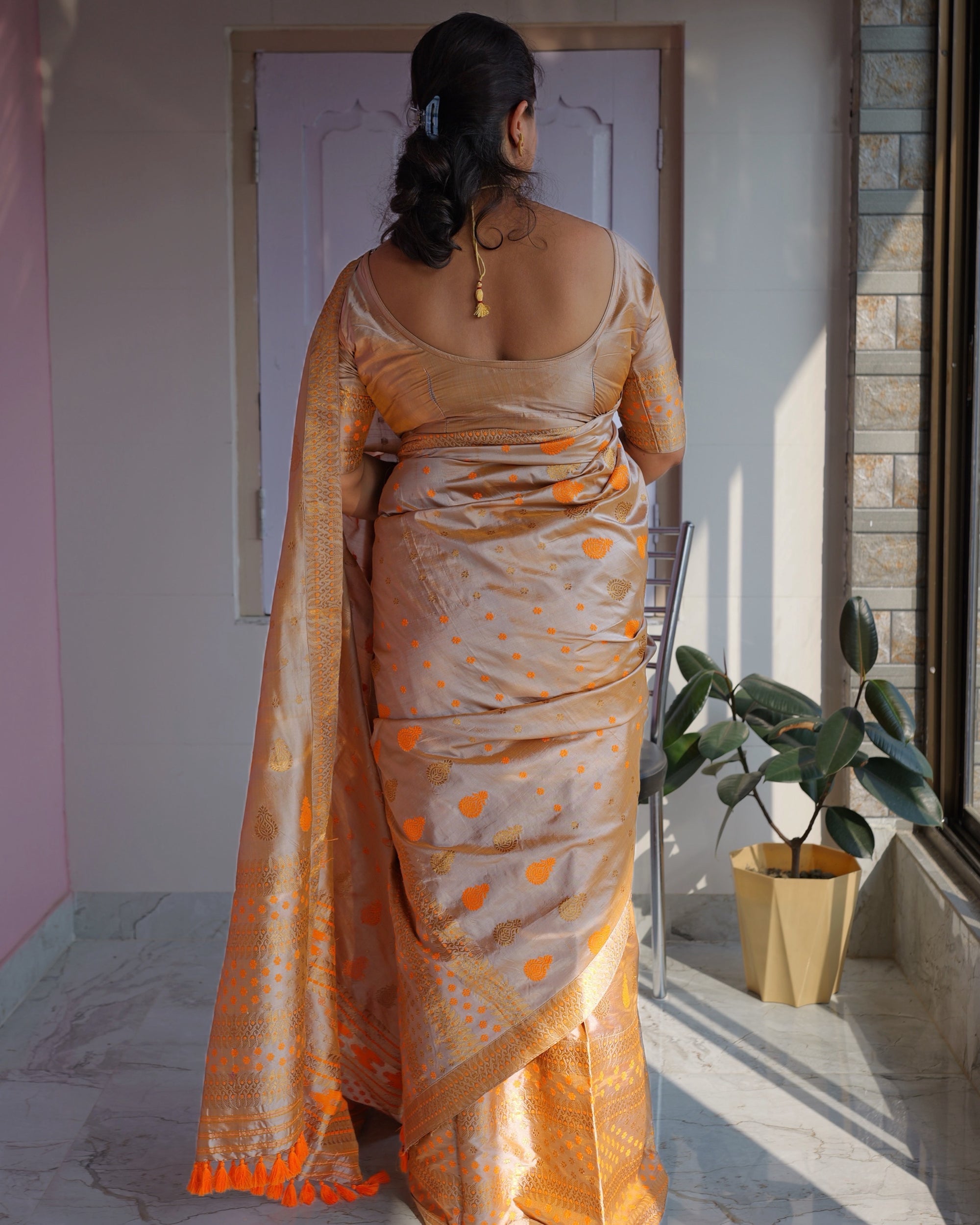 Seen from behind by a sunlit window, the whole set visible: the sador falling from the shoulder to saffron tassels at the floor, saffron and gold paisleys across the cloth and bands of zari at the hem.