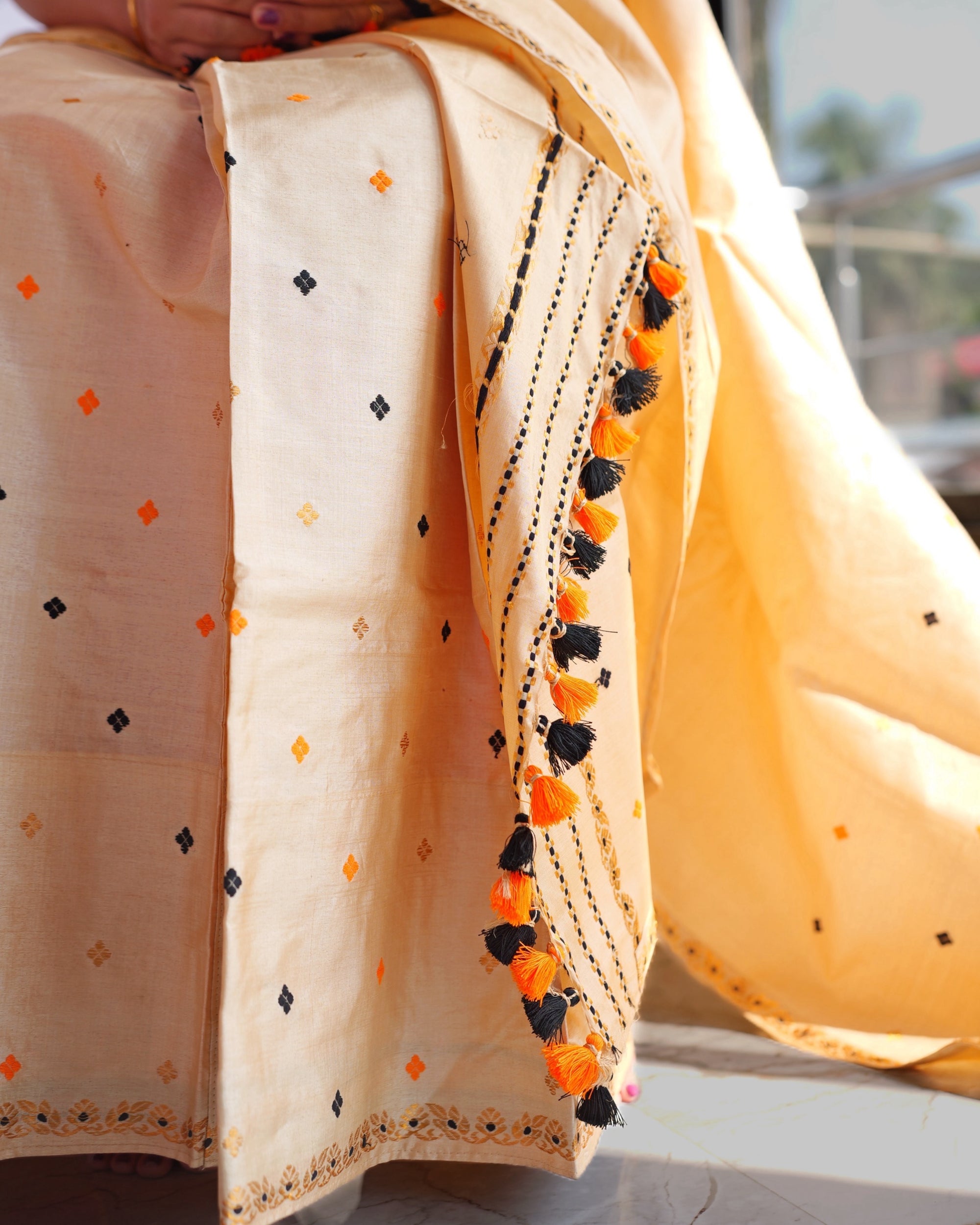 The lap close up. Small four-petal motifs in orange, black and gold across the cream body, the end of the sador hanging down in lines of tiny checks with orange and black tassels, and a narrow band of gold flowers at the hem.