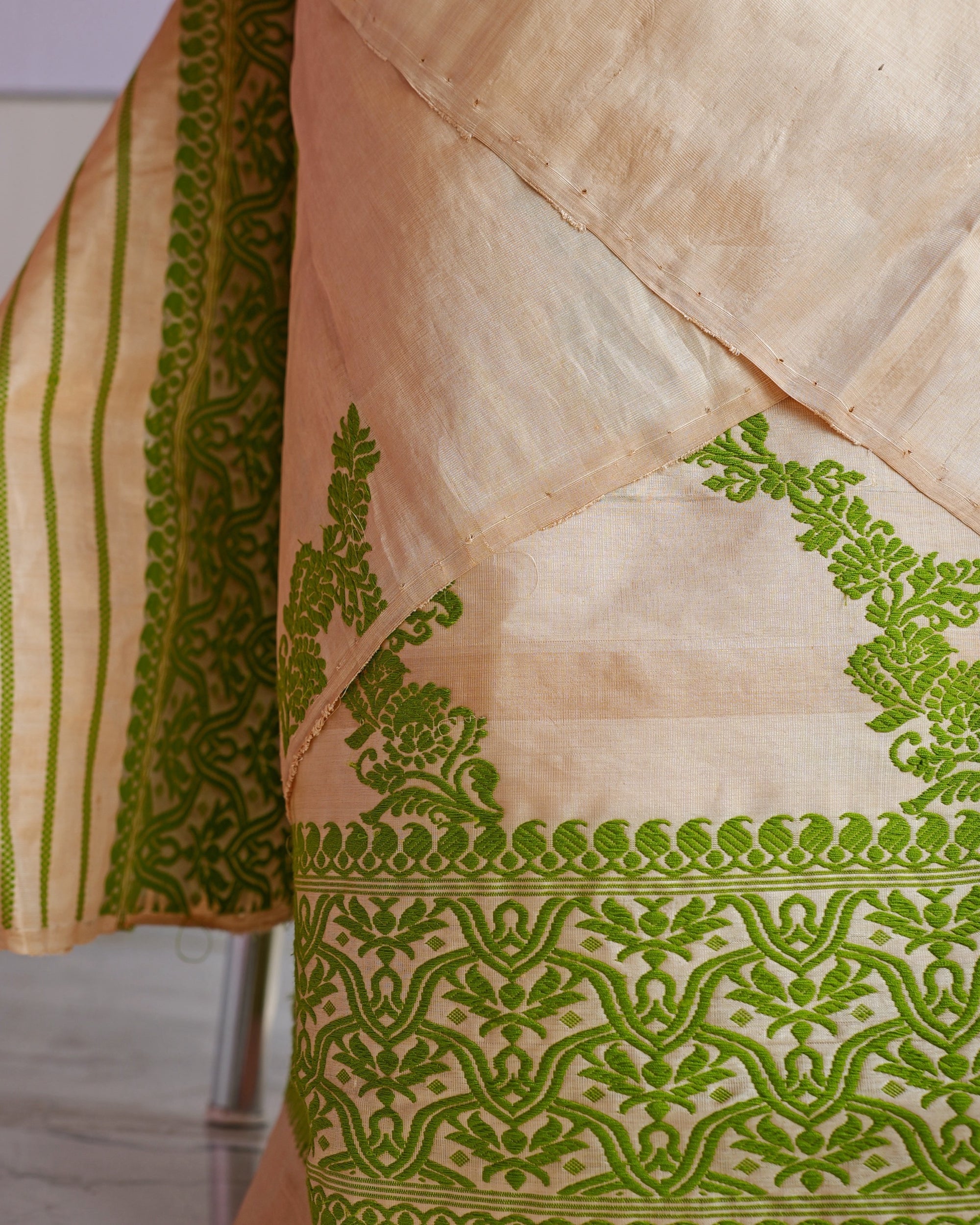 The hem of the mekhela close up. A wide green border of interlaced arches under a scalloped edge, leafy sprays rising above it, and the end of the sador hanging at the left in a band of green vines and stripes.