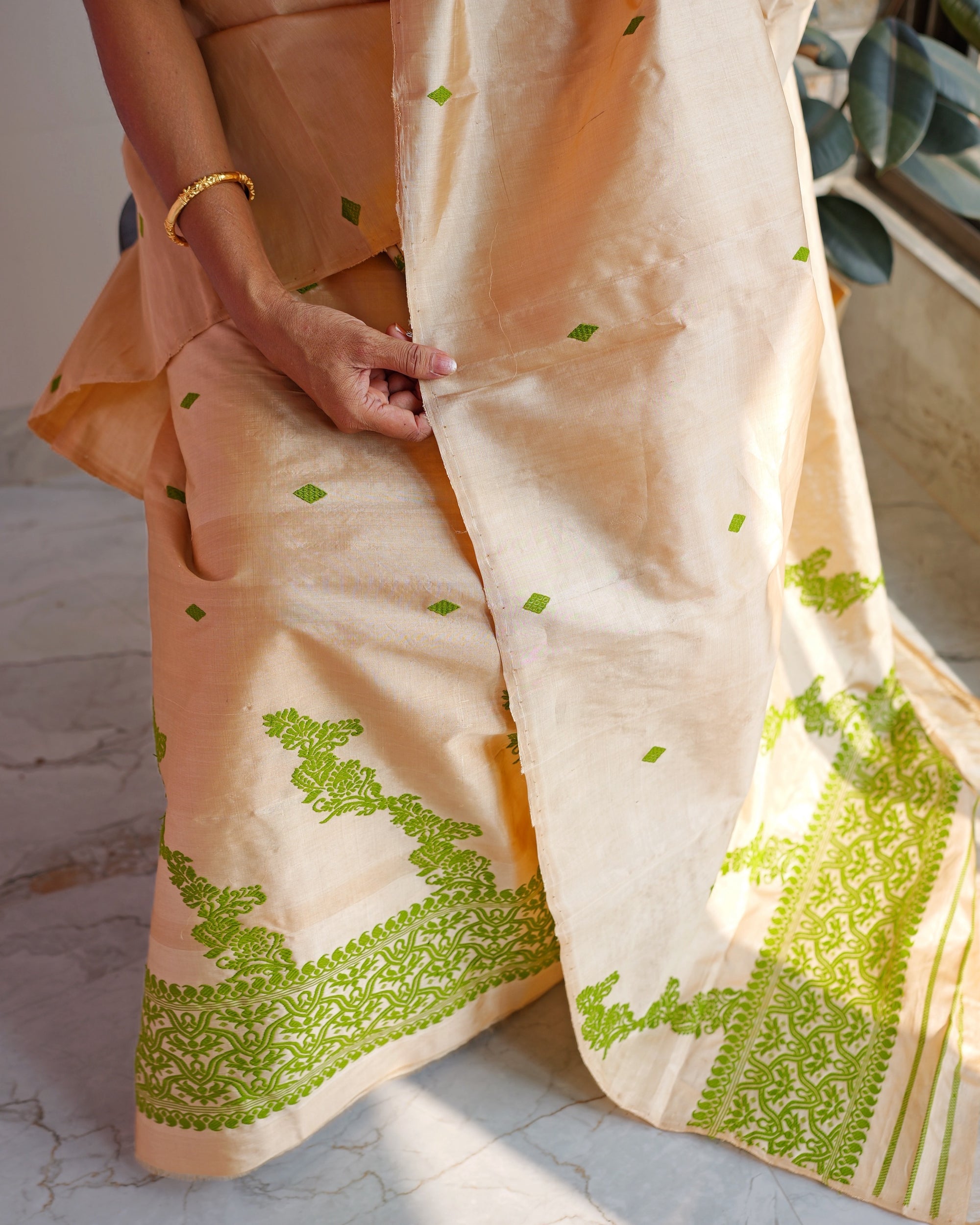 Seated, from the waist down, fingers pinching the edge of the sador. Small green diamonds across the golden-beige body, green scrollwork climbing in steps from the hem, and the green end panel of the sador at the right.