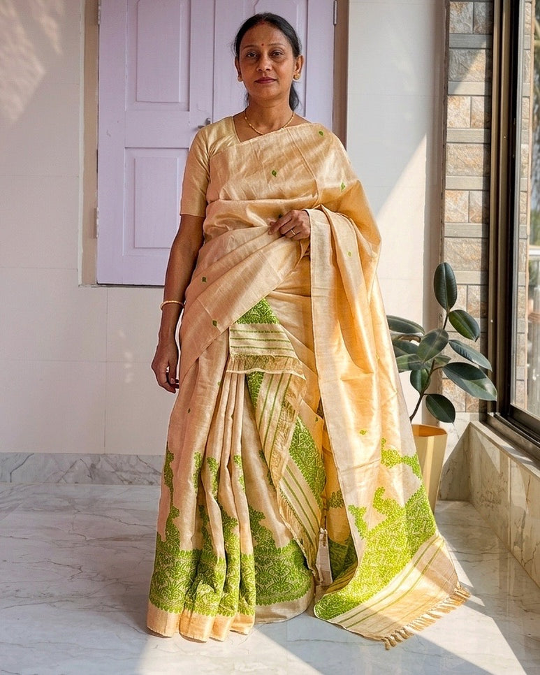 Standing against a white door, the whole set visible: a cream sador and mekhela with green vine borders and green striped panels at the hem.