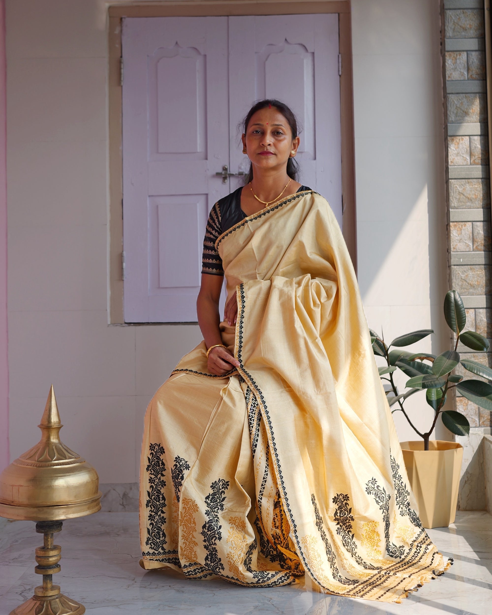 Seated against a lilac panelled door in a black blouse, the whole set visible to the floor. A golden-cream drape edged in black falls from the shoulder, and black and gold creepers climb the hem, beside a brass xorai and a potted rubber plant.