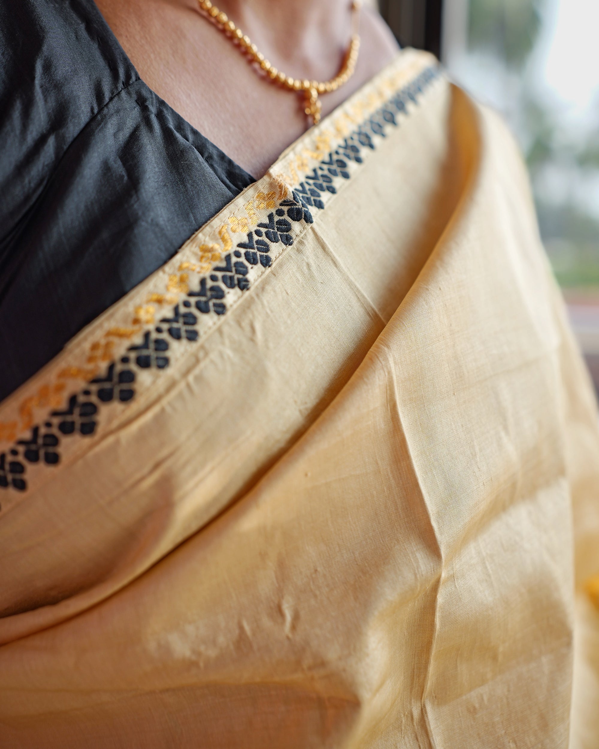 The border where it crosses the collarbone: small black four-petal florets in a row beside a wavy gold line on golden-cream silk, over a black blouse, with a gold bead necklace.
