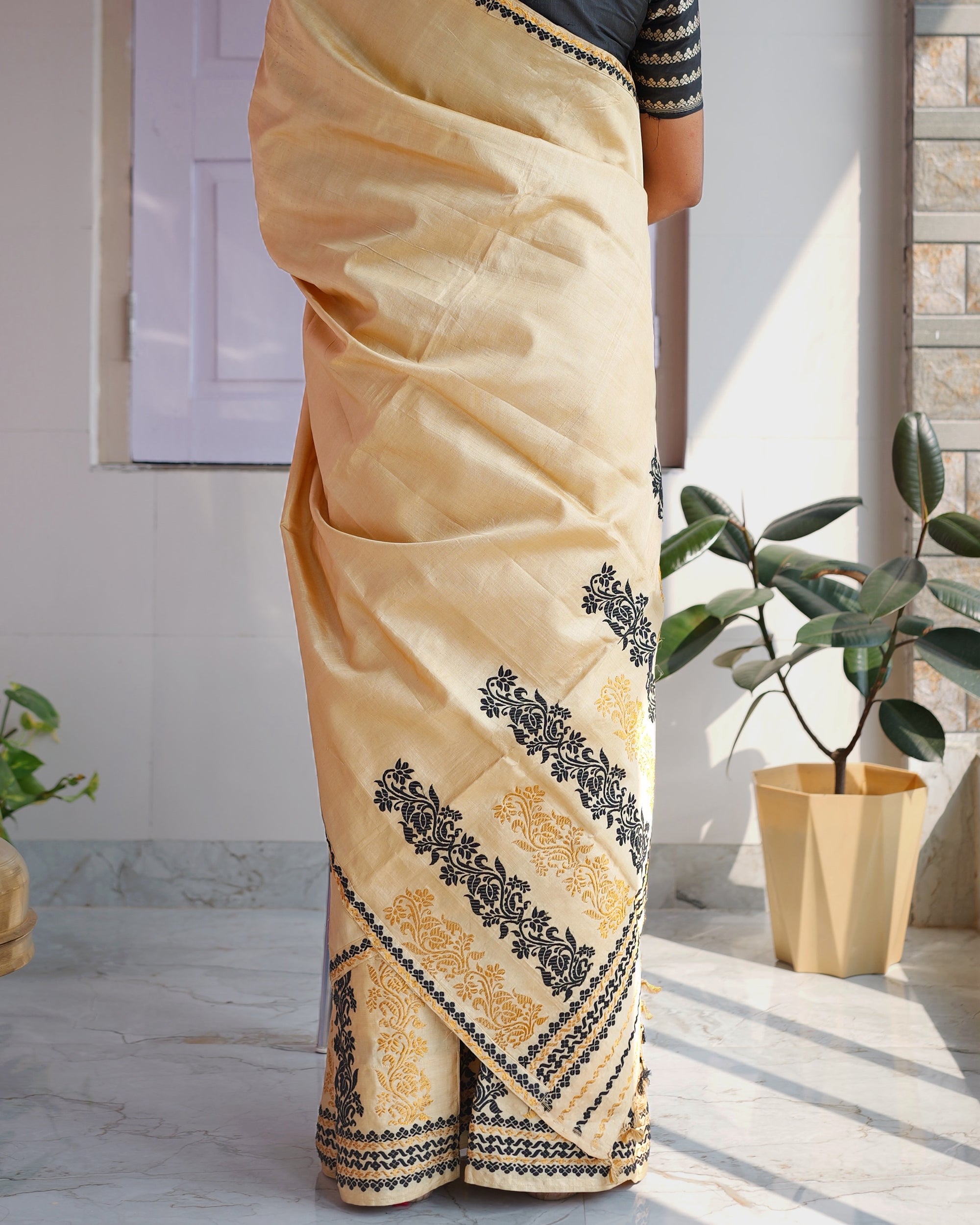 Standing, seen from behind and framed from the shoulders down. The drape falls down the back to the hem, its end carrying black and gold creepers and banded stripes, beside a rubber plant.