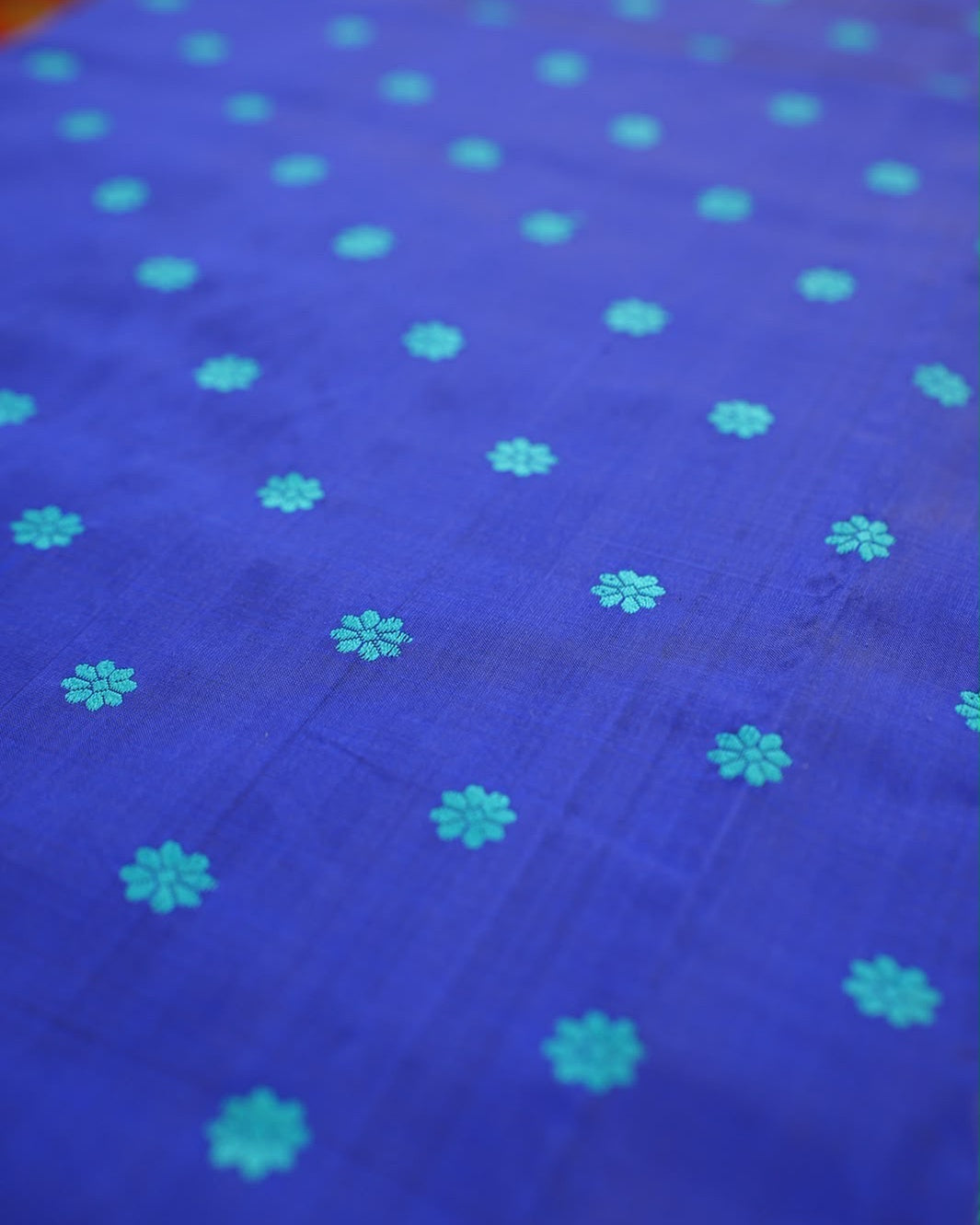 The body close up, laid flat and in shallow focus: small eight-petal turquoise flowers in spaced rows on royal blue, with faint lines of the weave in the ground.