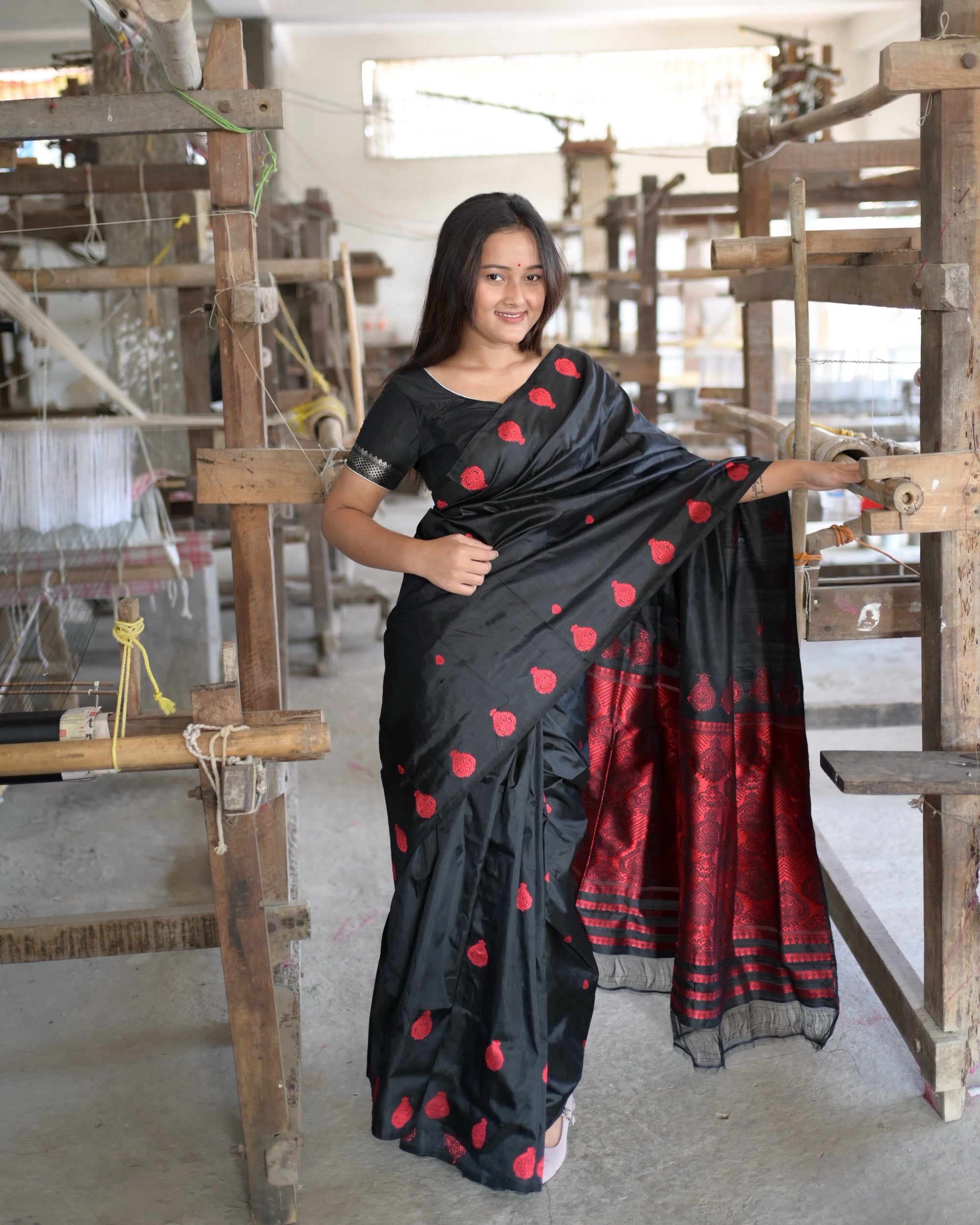 Holding the pallu out at arm's length beside a loom: red teardrop medallions on black, then the pallu's red field and red stripes at its end.