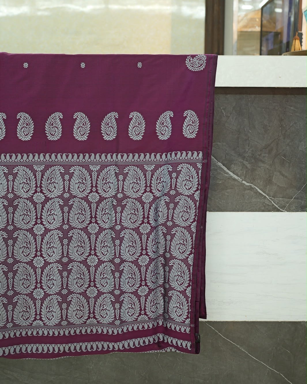 The pallu hung over a grey stone counter: small flowers spaced across a deep wine body, a row of large silver-white paisleys, then a field of paisleys and round flowers framed by bands of small curled paisleys, one above and two below.