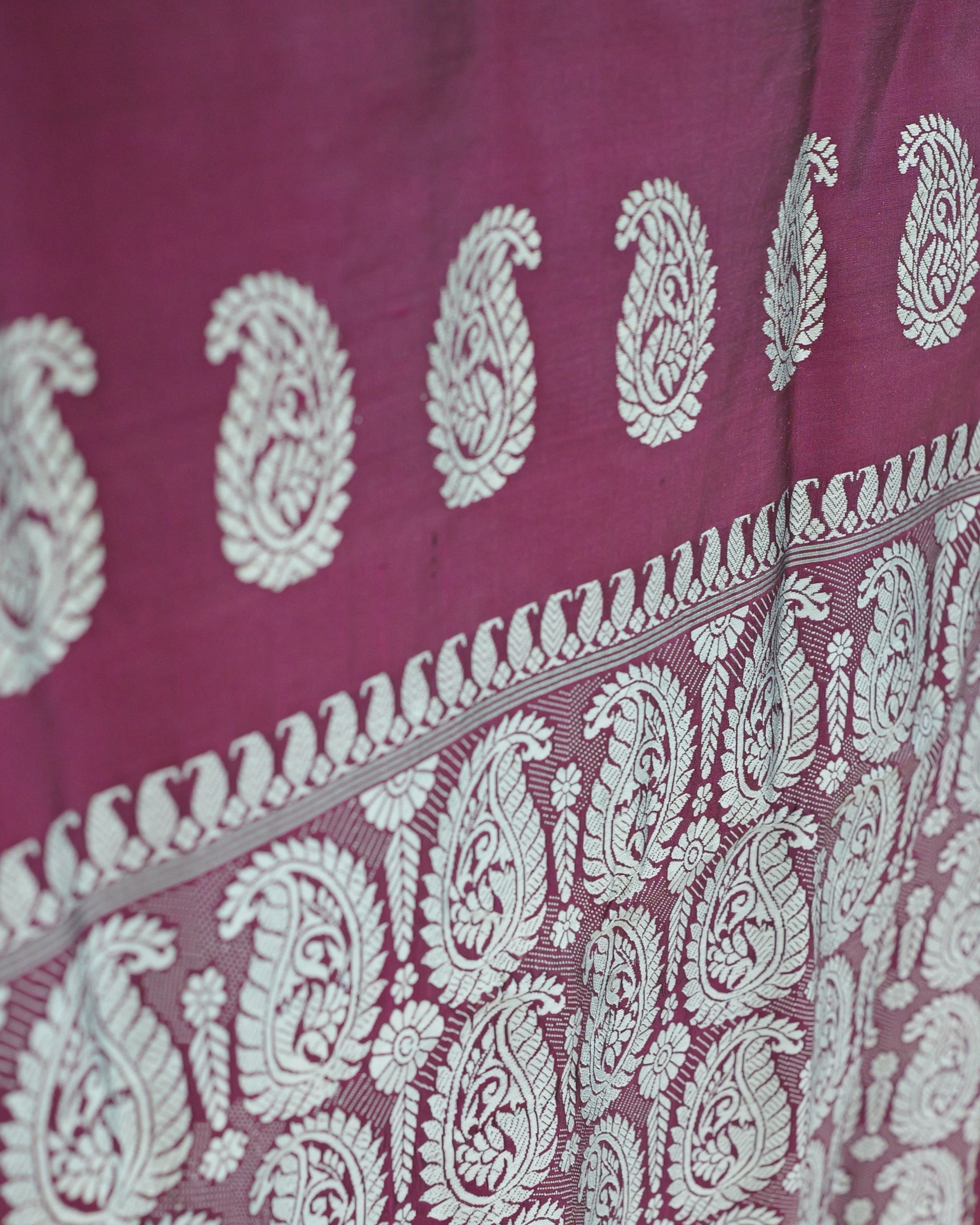 Close up where the body meets the pallu: large silver-white paisleys filled with leaf and vine work, a band of small curled paisleys and fine stripes, and the start of the paisley field below.
