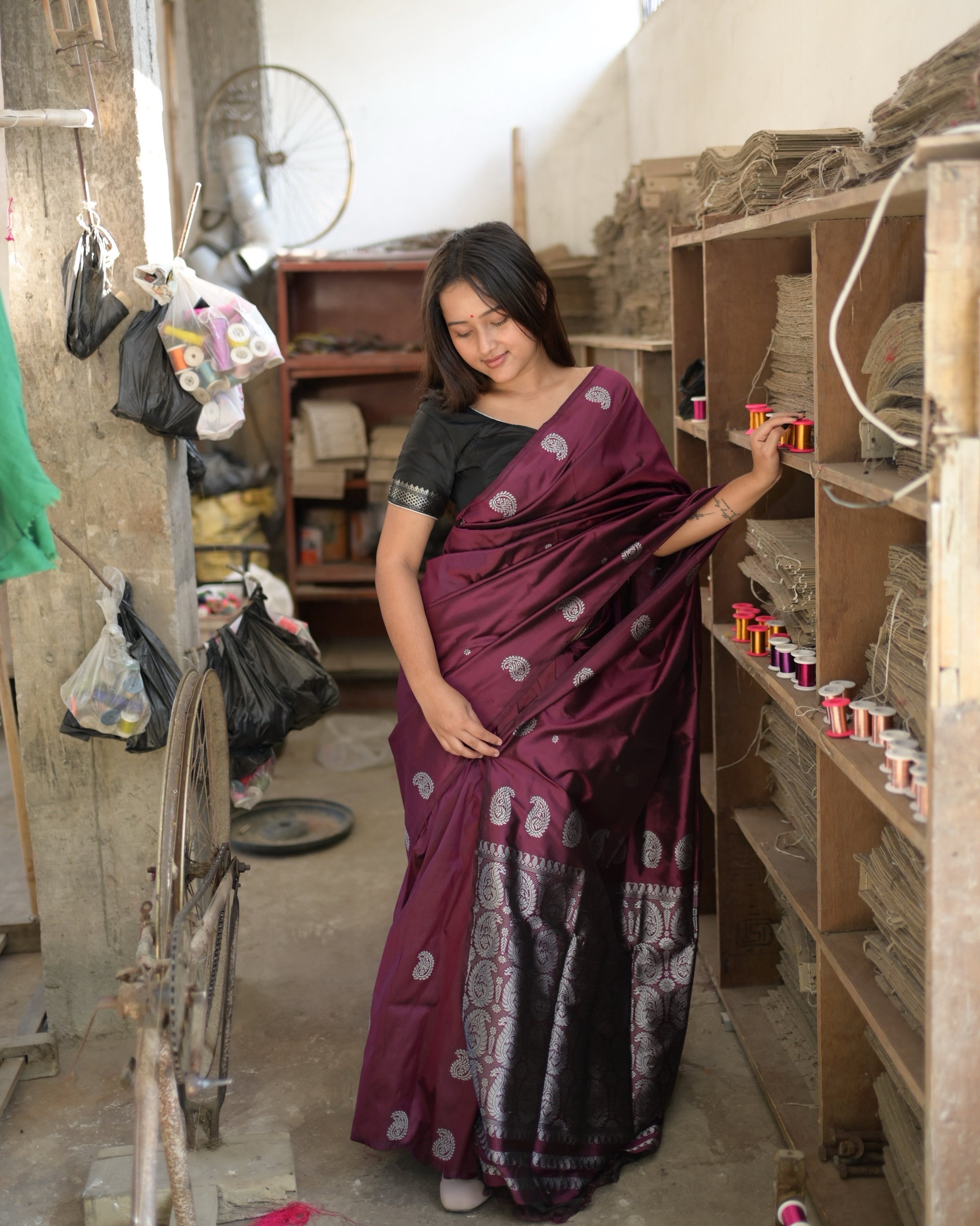 Eyes lowered, one hand on a shelf of thread reels: silver-white paisleys on wine, and the paisley-filled end of the saree at the hem.