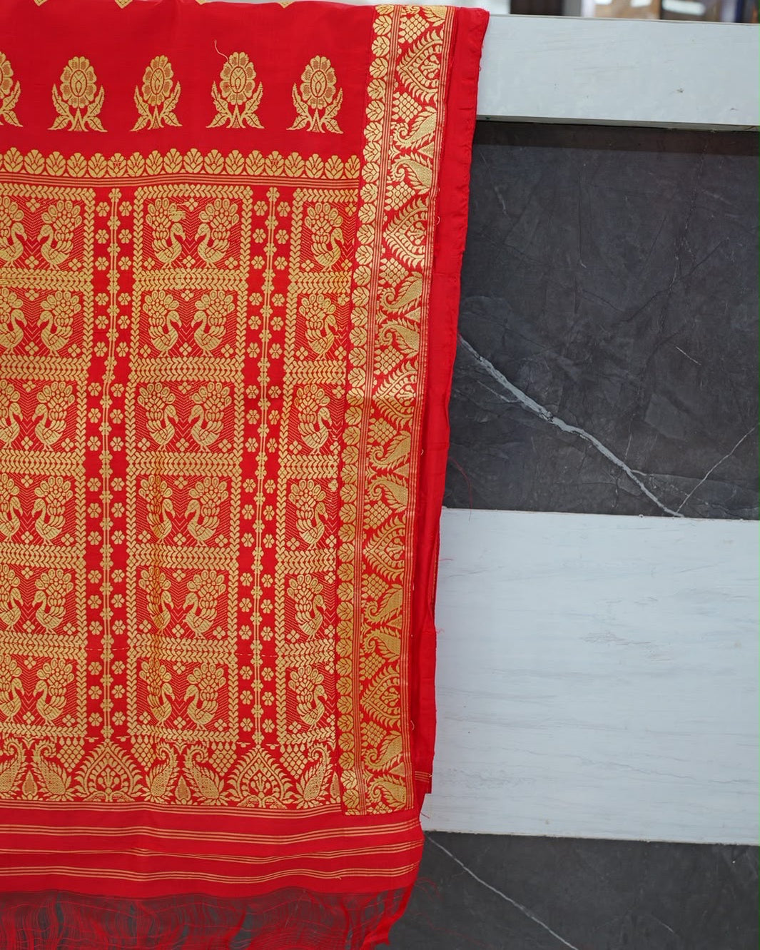 The pallu hung over a stone counter and seen straight on: a row of golden flower sprigs on red, a grid of panels each holding two birds facing each other, then golden stripes and a loose red fringe at the foot, with a broad golden border down the right side.