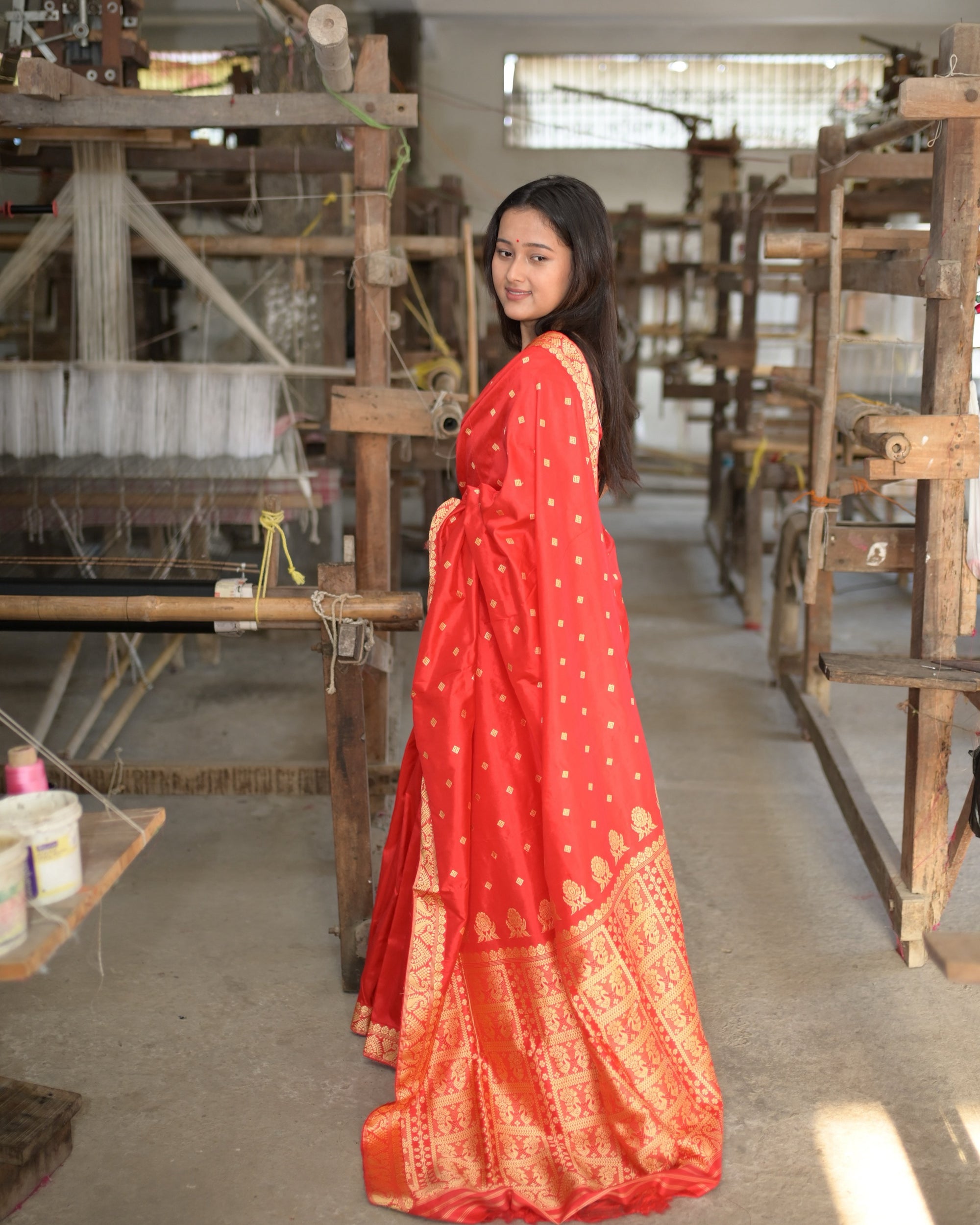 In profile among the looms, looking back: the red pallu down the back, worked in gold from the waist to the floor.