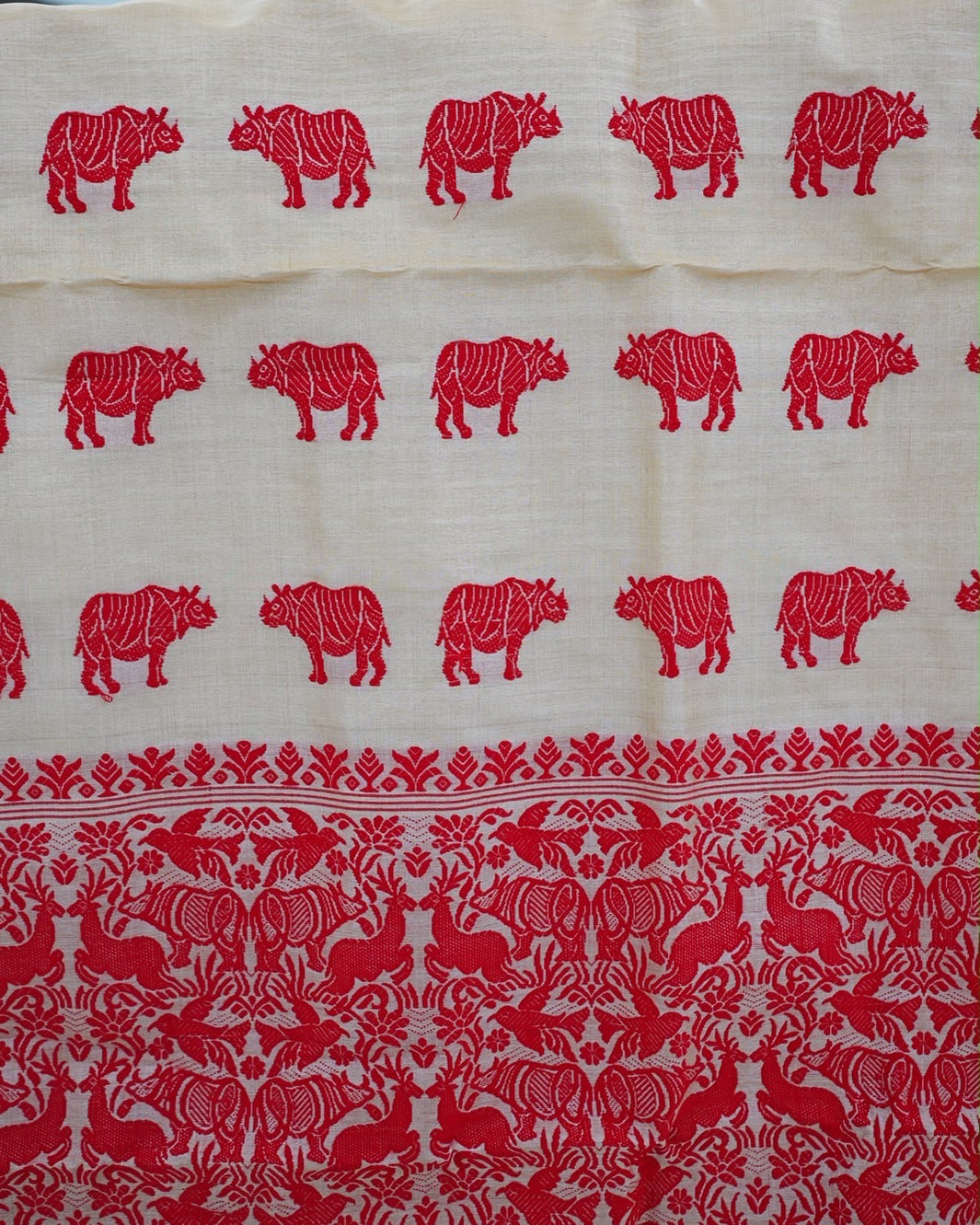 The saree hung up and seen close: three rows of red rhinoceros on cream, in facing pairs, above the pallu's red field of rhinoceros, deer and birds.