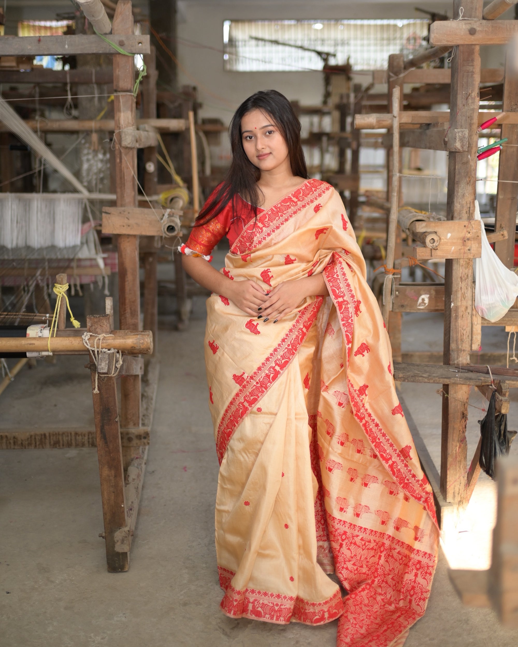 Standing between wooden handlooms in a weaving hall, in a red blouse, hands resting at the waist: small red rhinoceros across the cream body, the red border down the front and the pleats falling to the floor.