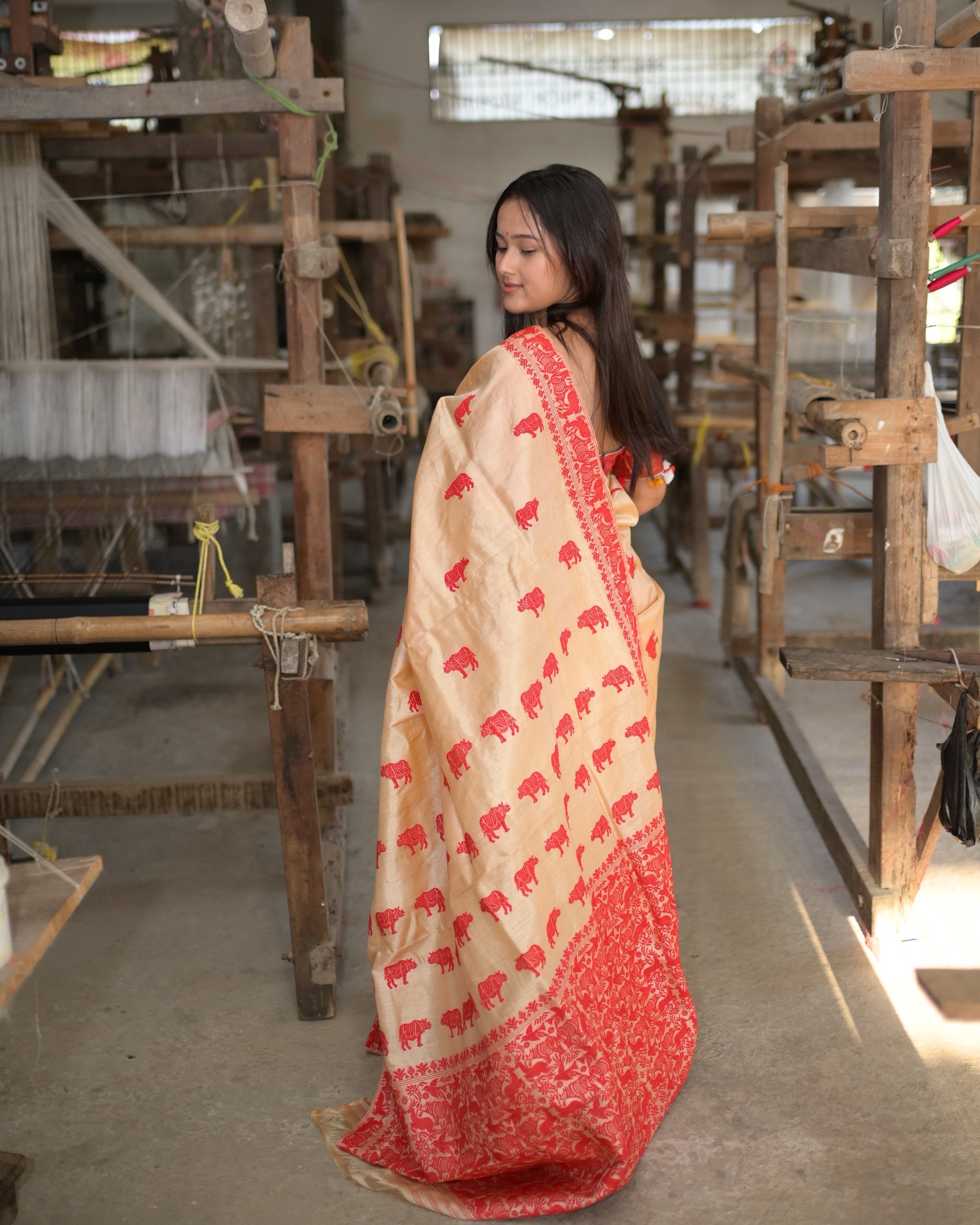From behind in the weaving hall, looking back over the shoulder: the pallu down the length of the back, rows of red rhinoceros on cream above its deep red forest panel.