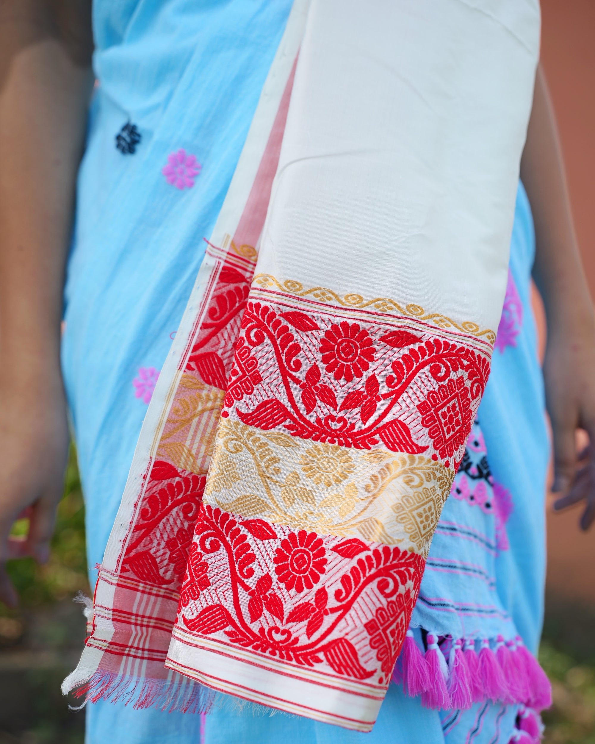 Hanging down in front of a pale-blue sador with pink tassels, the wearer's hands at her sides, in daylight: a plain ivory length ending in a slim gold scroll border and three bands, red, gold and red, each worked with a flowering creeper. At the corner, red side stripes and a short fringe of white and red threads.
