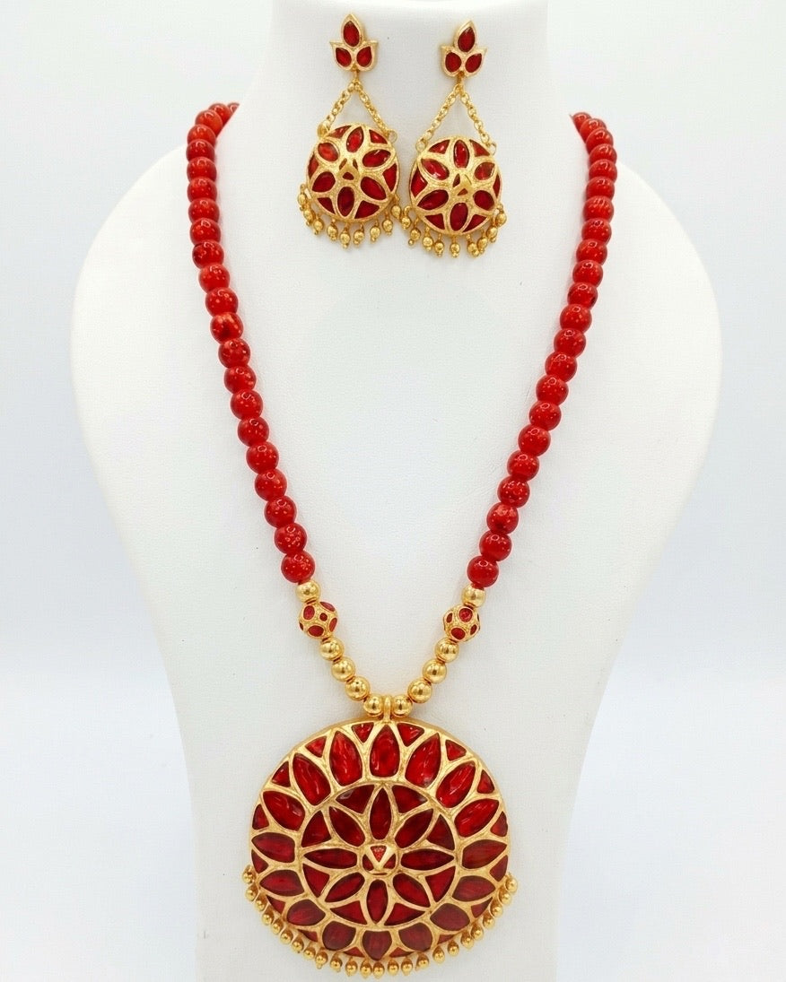 On a white display bust against a white ground: a large round gold medallion filled with red petal shapes in two rings, fringed with small gold beads along its lower edge, on a strand of round red beads finished with gold beads near the pendant. The earrings are smaller red discs hung from three-stone buds on fine chains.
