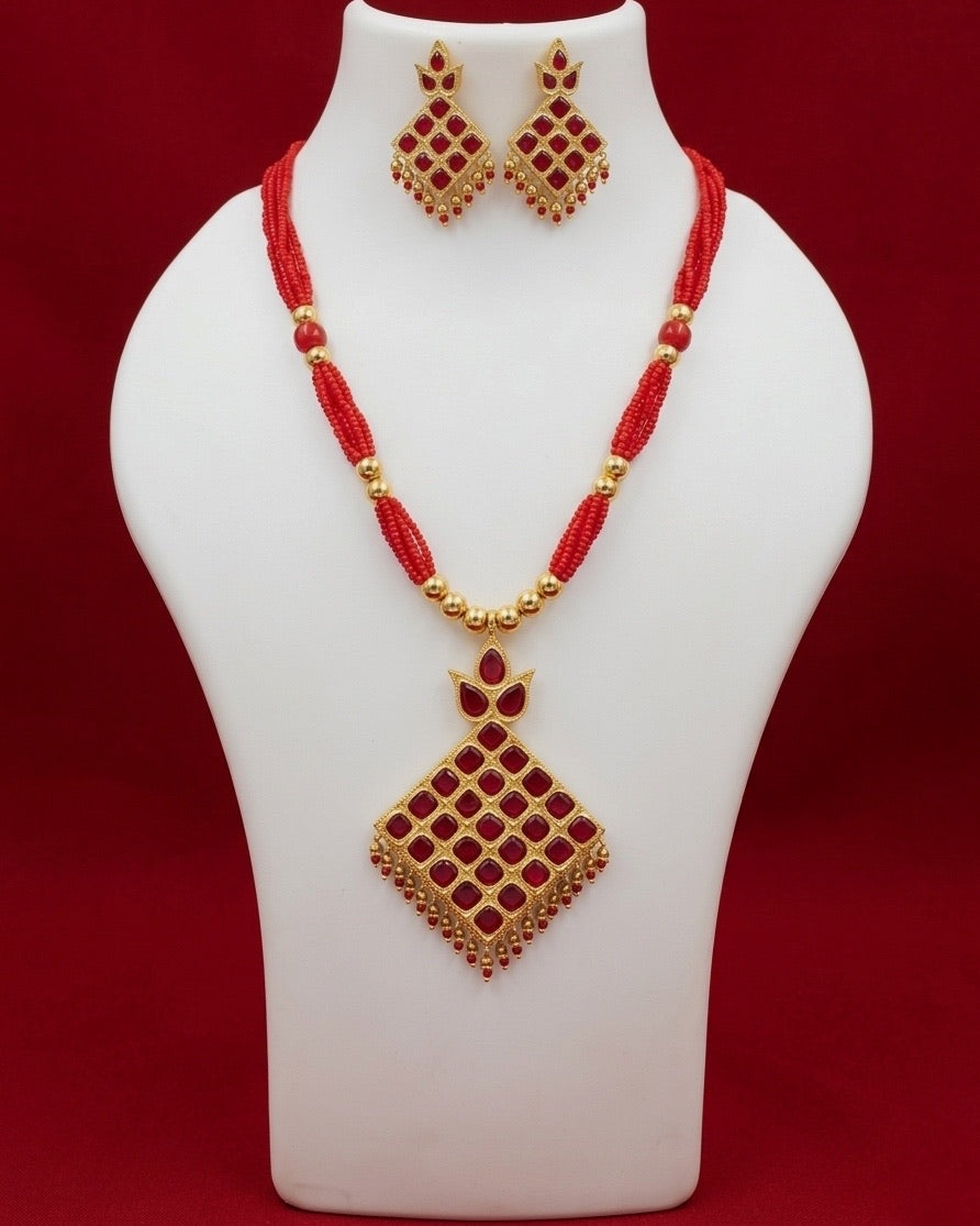 On a white display bust against a deep red backdrop: a gold rhombus set with square red stones on the diagonal, under a three-stone crown and fringed with gold and red bead drops, hanging from twisted strands of bright red seed beads gathered with gold beads. The earrings are the same rhombus, smaller.