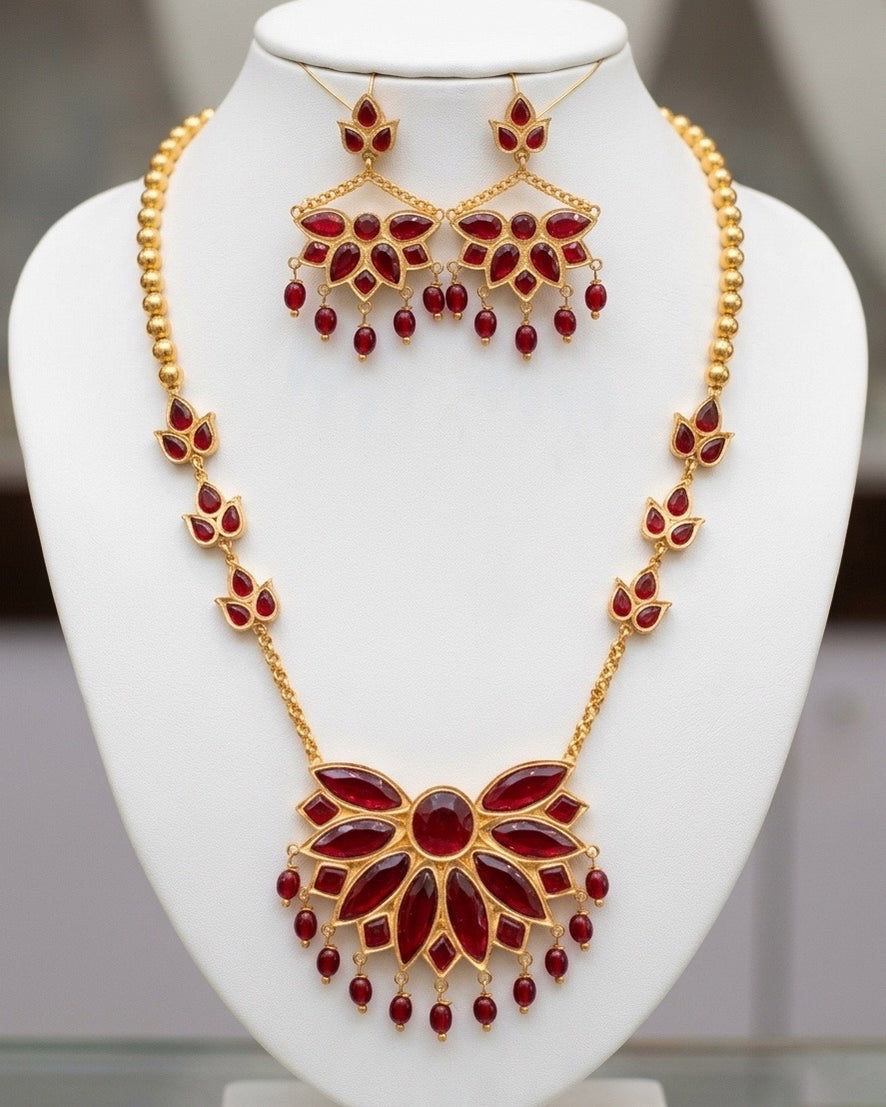 On a white display bust in front of a softly blurred grey room: a half-round fan of long red marquise stones around an oval centre, fringed with red bead drops on gold, on a chain of gold beads and three-stone links. The earrings hang from fine gold wires, each a smaller fan below a three-stone stud and a swag of gold chain.