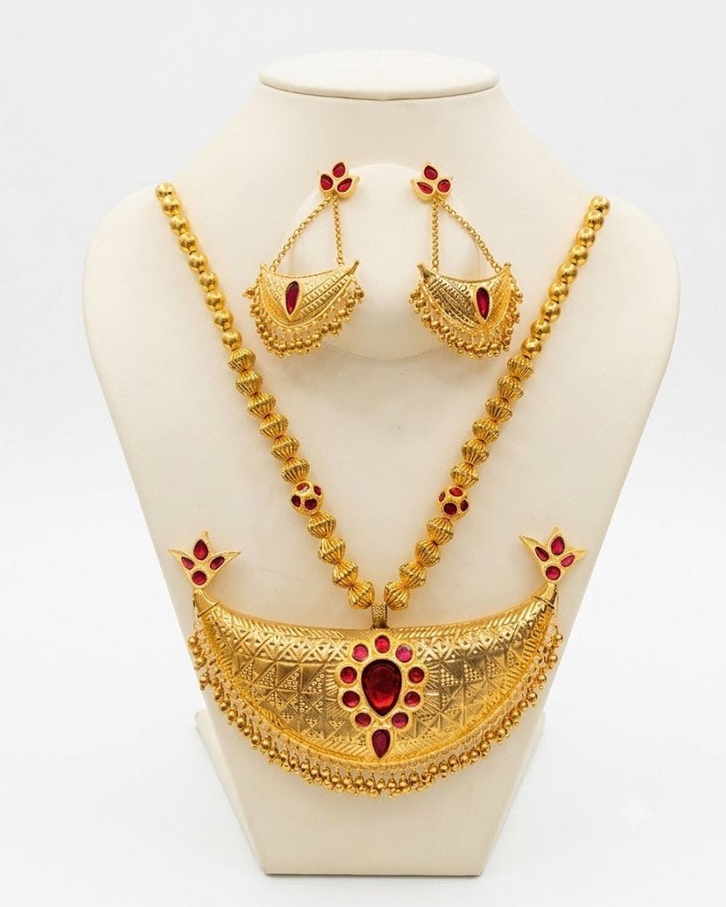 The necklace and earrings on a cream display bust against white. A broad crescent engraved in rows of dotted triangles holds a large red drop in a ring of small red stones, with a four-stone bud at each tip and a fringe of small gold drops, on a chain of plain and ribbed gold-toned beads. Crescent earrings, each with one red stone, hang on fine chains above.