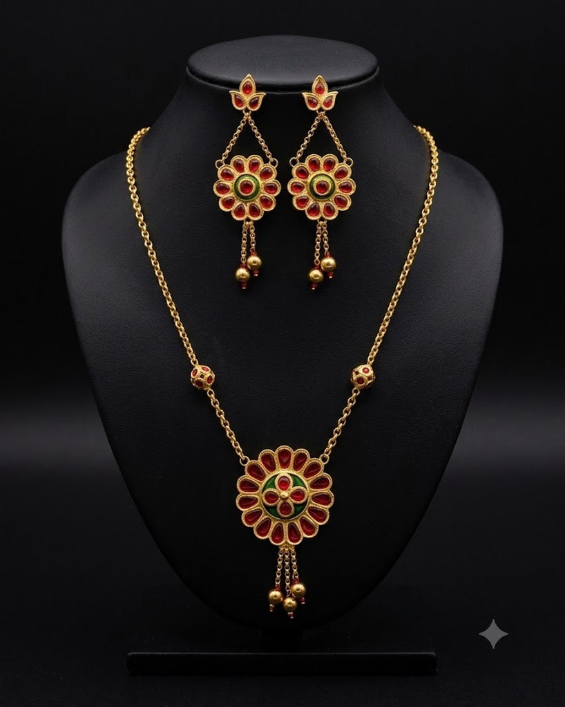 The necklace and earrings on a black display bust against black. A round flower of sixteen red petals around a green ring hangs from a fine gold chain, three short gold-ball drops below it and a red-set bead on the chain at each side. The earrings are smaller flowers on fine chains from three-stone studs, each ending in two gold-ball drops.