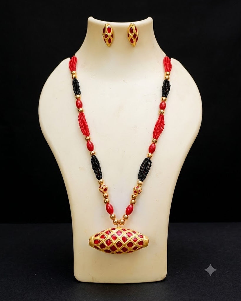 The necklace and earrings on an ivory display bust against black. A barrel-shaped pendant lies on its side under a diagonal gold lattice, red in every cell, hung from a strand of red and black seed-bead bunches, red oval beads and small gold beads. Two small studs with the same red lattice sit at the neck above.