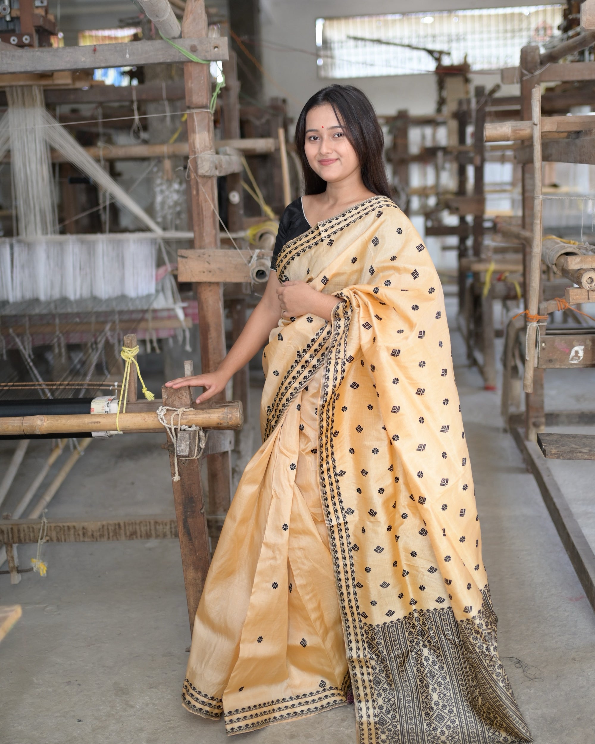 Standing at a wooden handloom in a weaving hall, one hand on its beam, smiling: the golden beige saree full length, small black flowers and squares across the body and the black pallu panel at the hem.