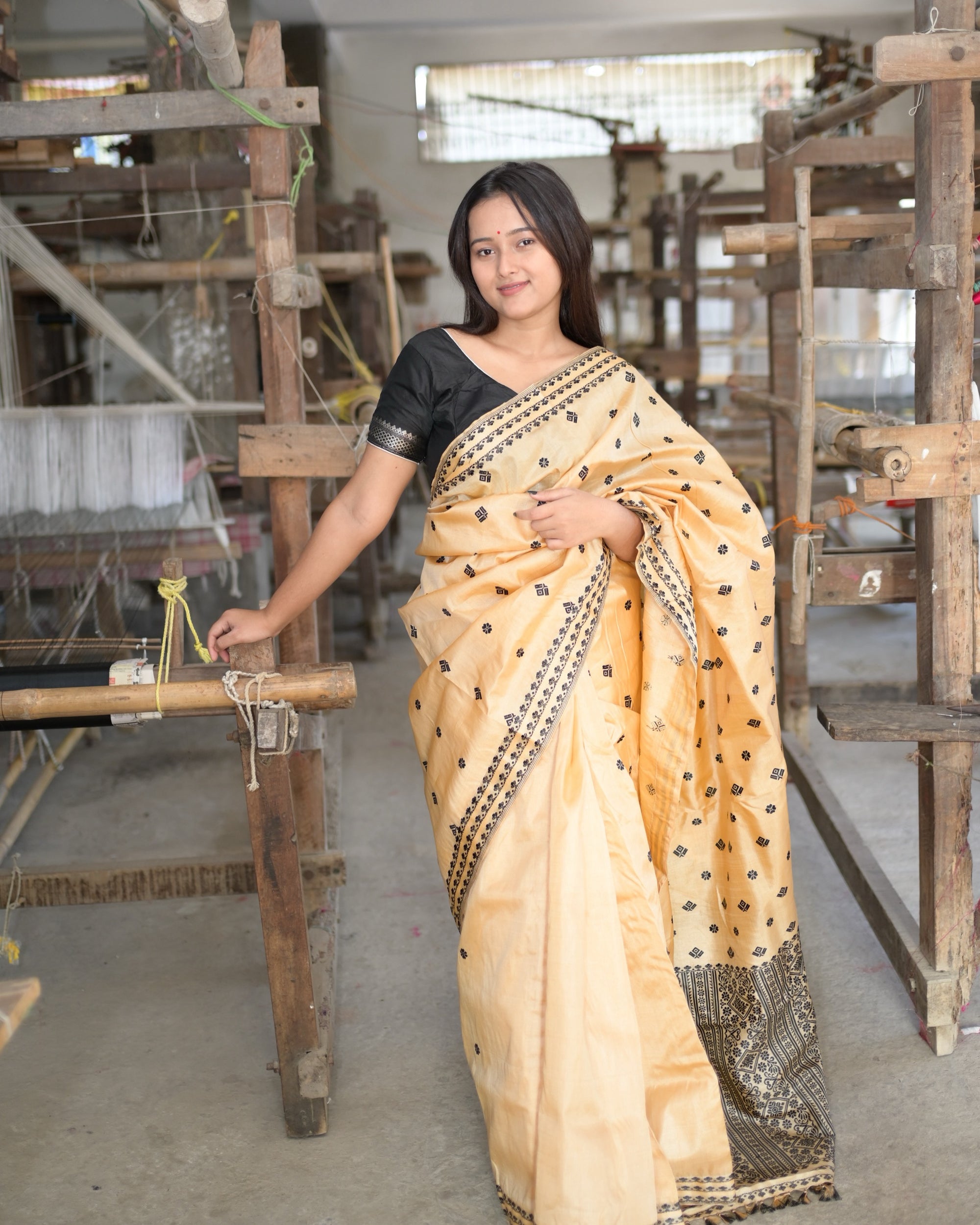In a black blouse, one hand on a handloom, the pallu gathered across the front: black flowers and squares on golden beige and the floret border along the edge.
