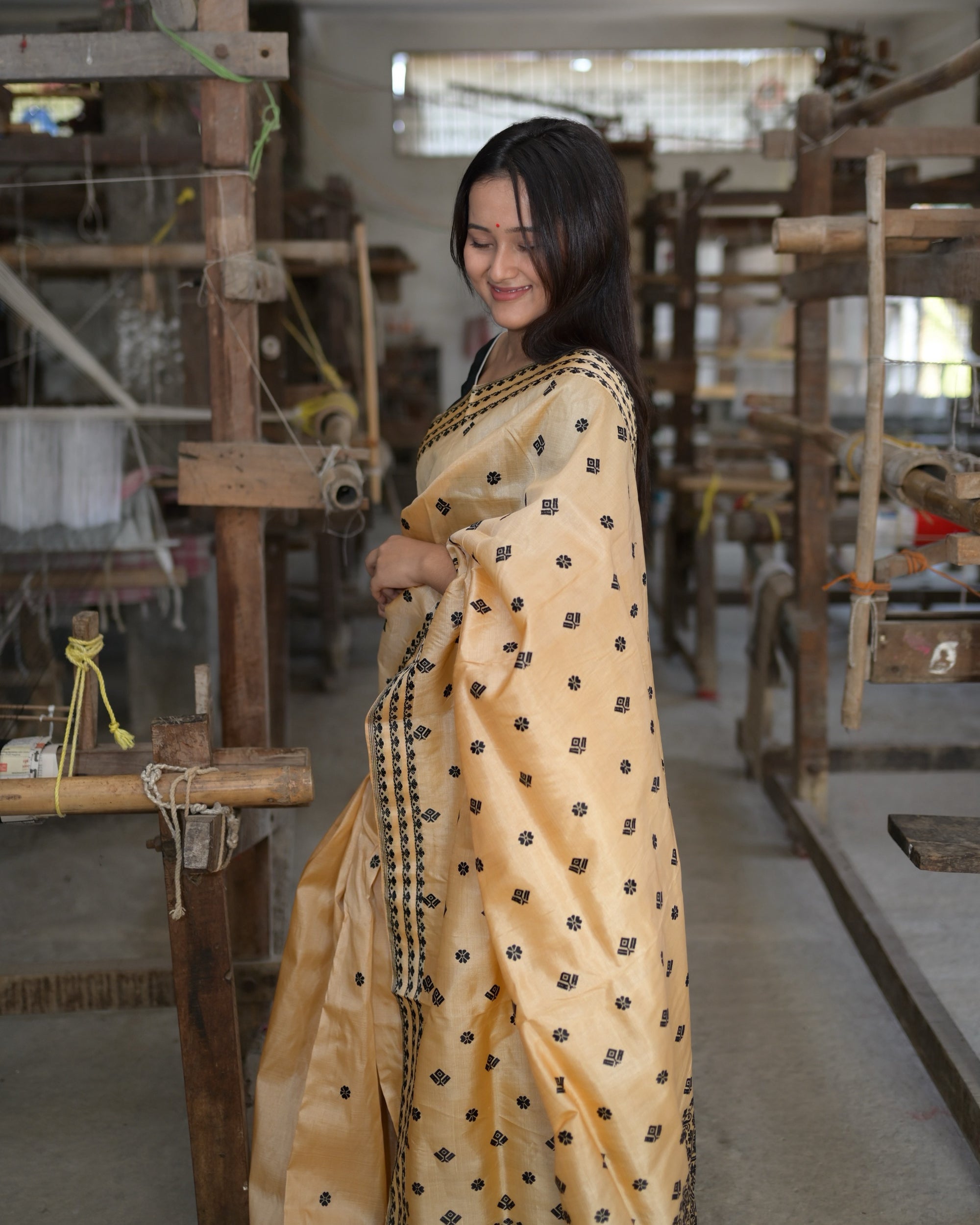 Eyes lowered between the handlooms, the pallu drawn across the body: black flowers and squares spaced over the golden silk, the floret border down the front.