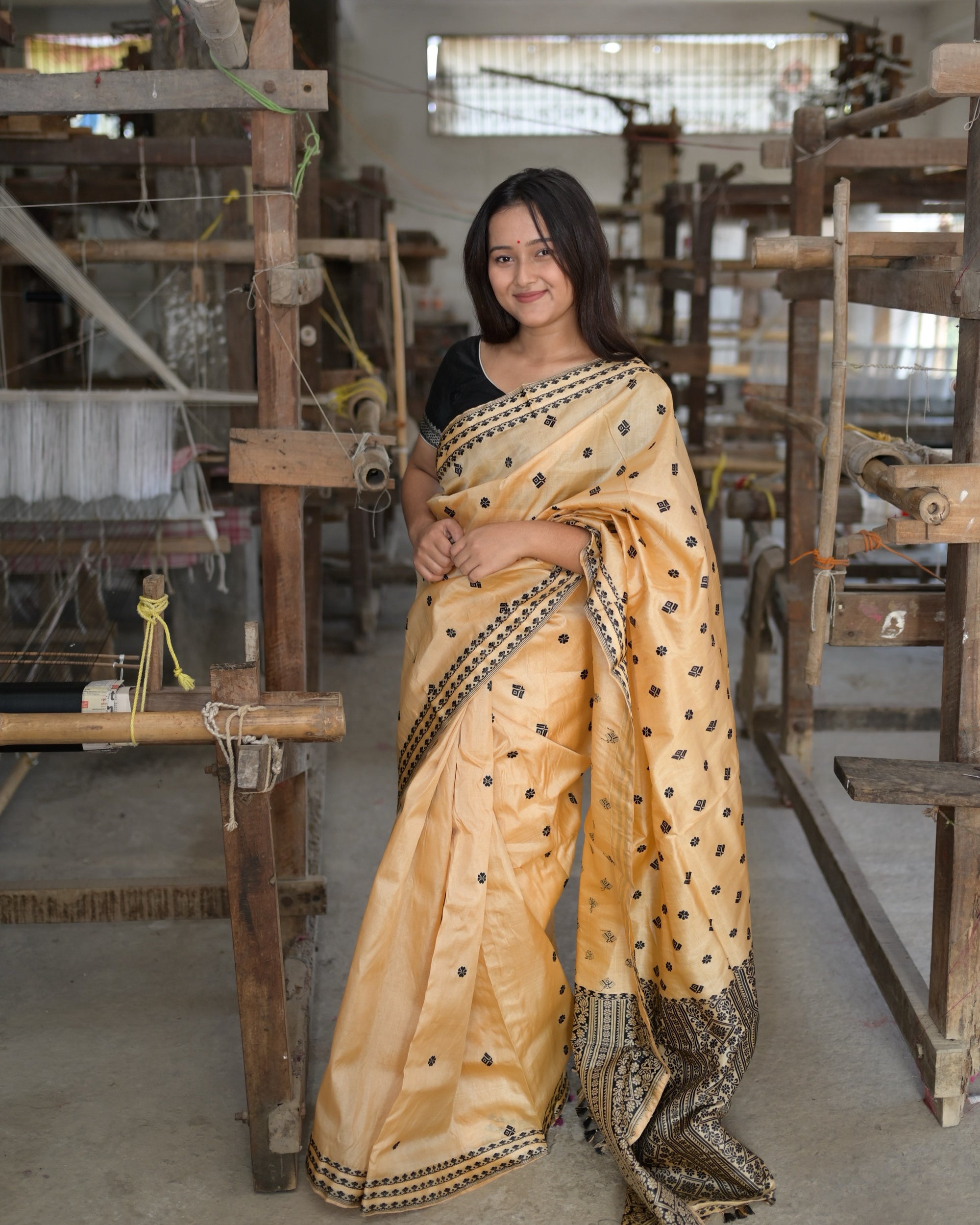 Full length among the looms, a hand at the waist, smiling: the golden body with its black buti above the dense black pallu at the floor.