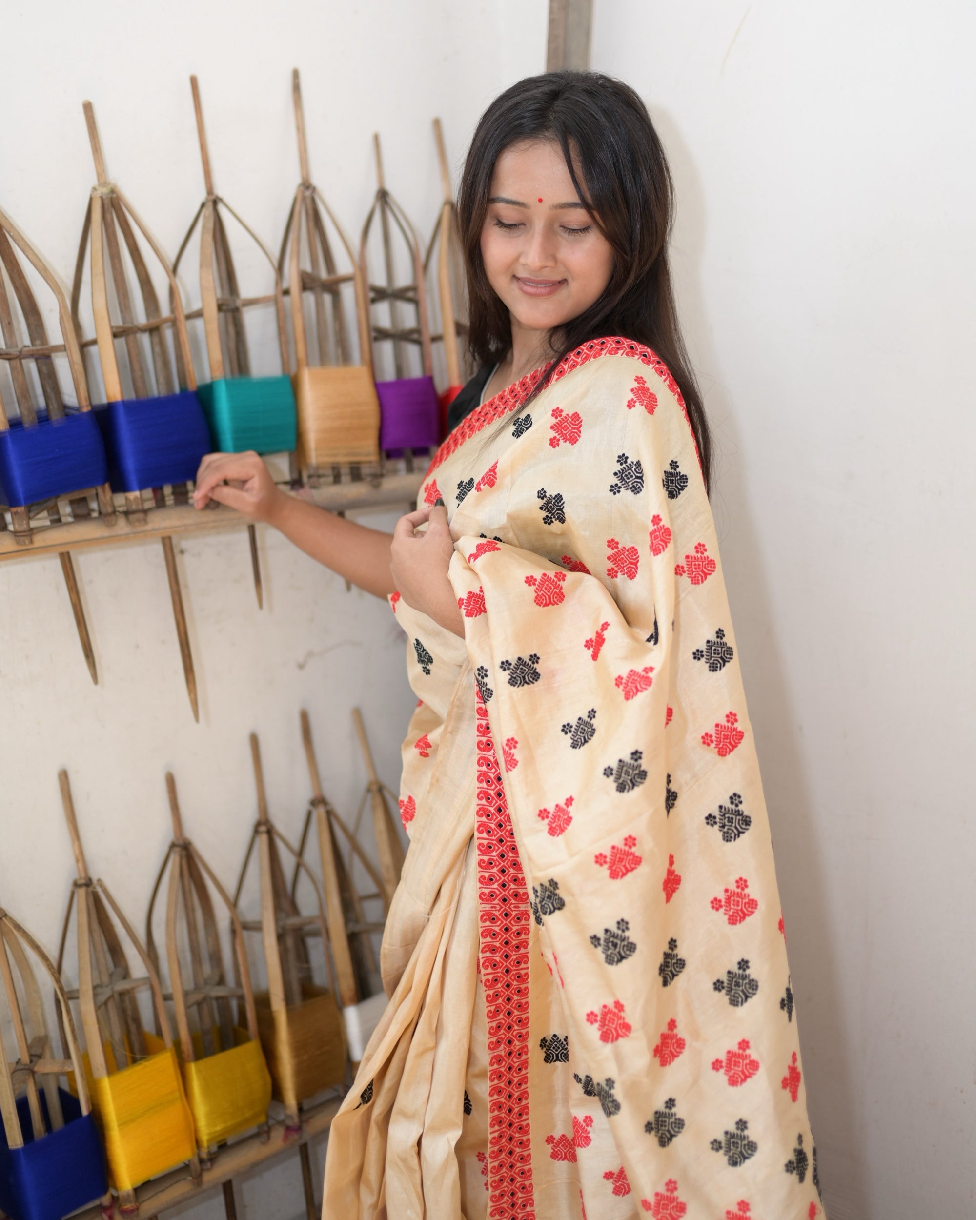 Closer, smiling, one hand on the reel shelf: the pallu gathered over the shoulder, small stepped buti in red and black on cream, and the red scroll border along its edge.