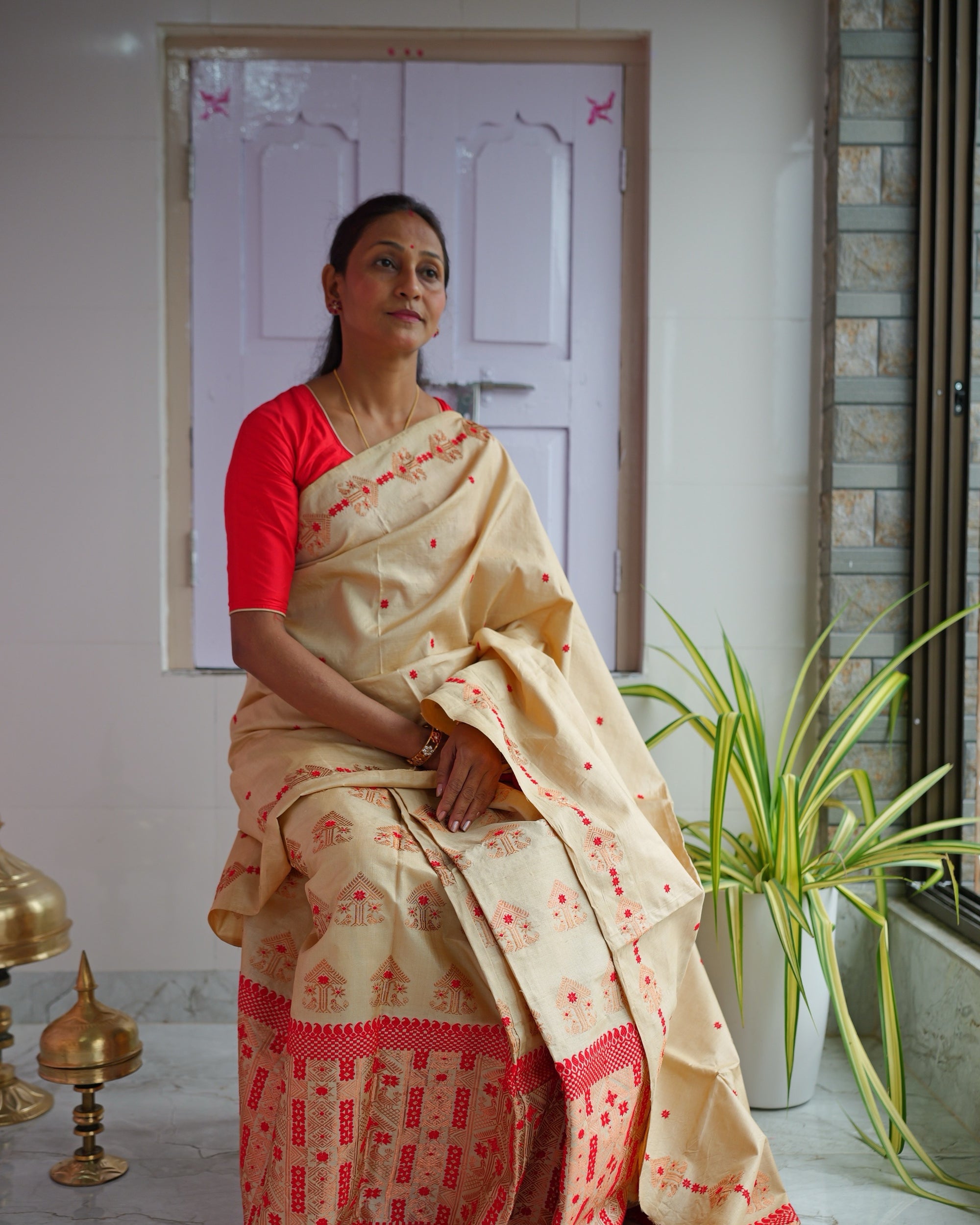 Deepa Kakoty seated against a lilac panelled door, in a golden-beige mekhela sador patterned in red and a red blouse, hands folded in her lap, looking away to one side.